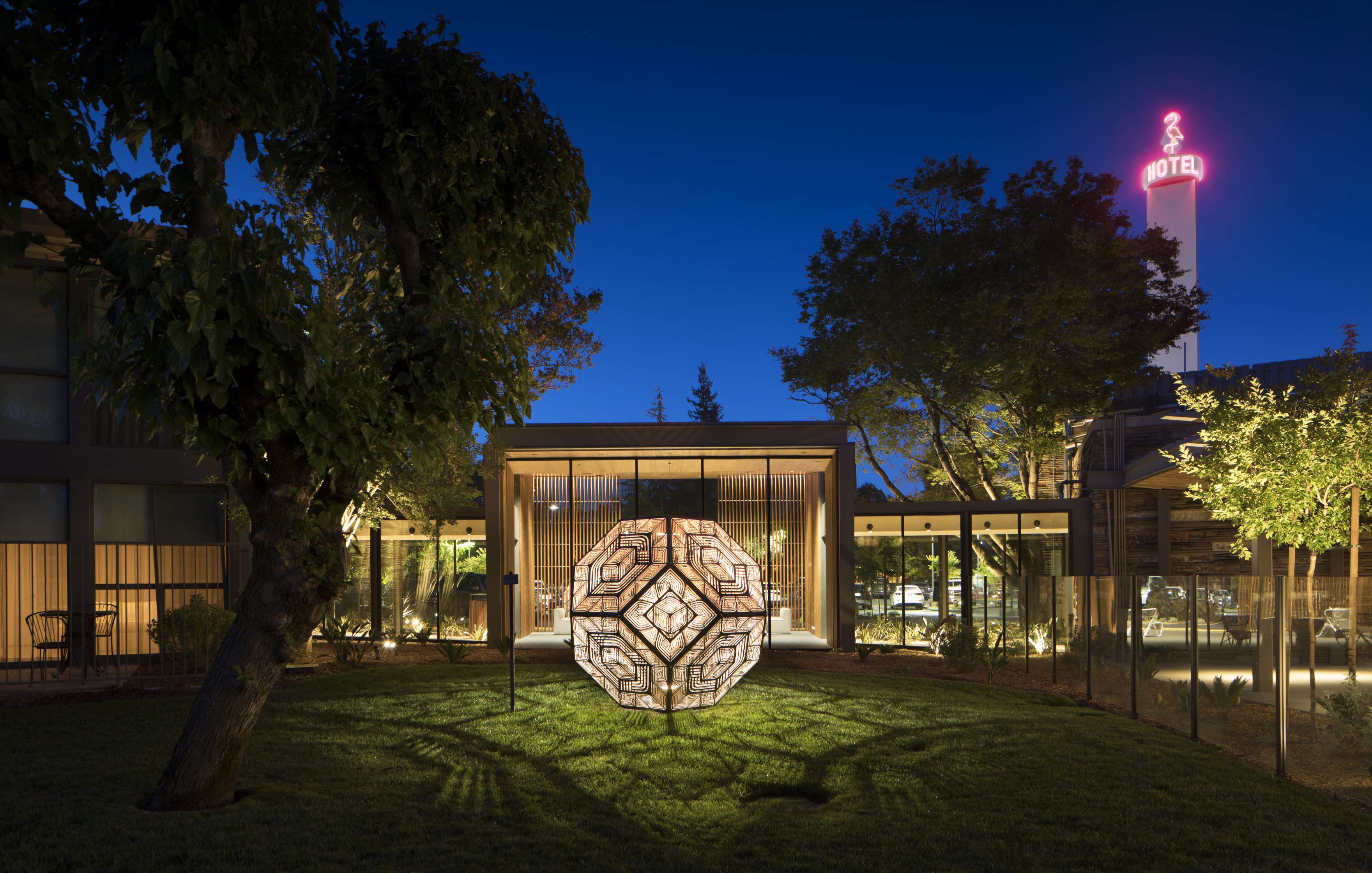 A geometric light sculpture is illuminated at night in a grassy courtyard, with modern buildings and a glowing motel sign in the background. Trees surround the scene.