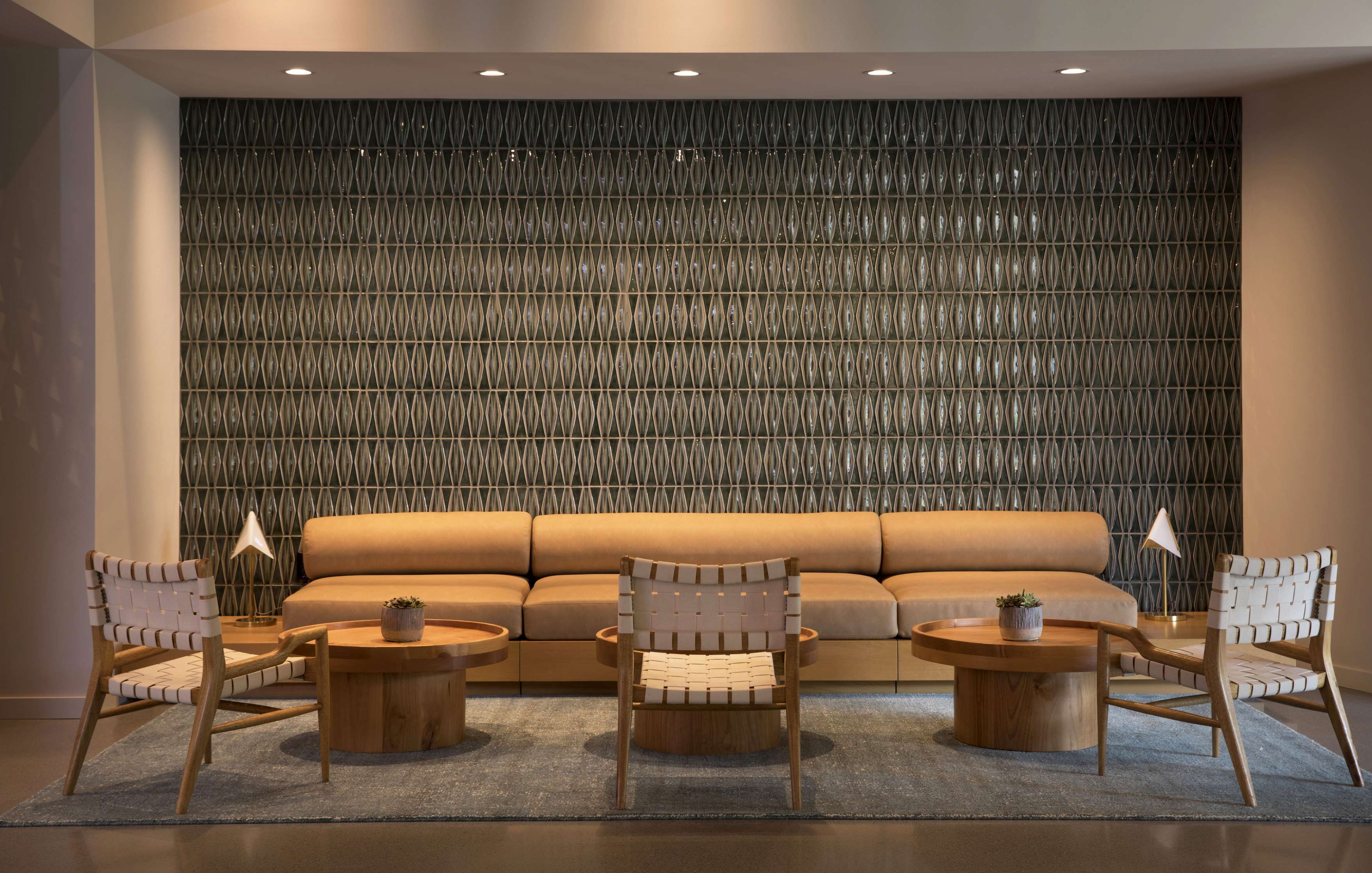 A contemporary lounge area with four wooden chairs, three round tables, a long sofa, two small lamps, and a patterned accent wall.