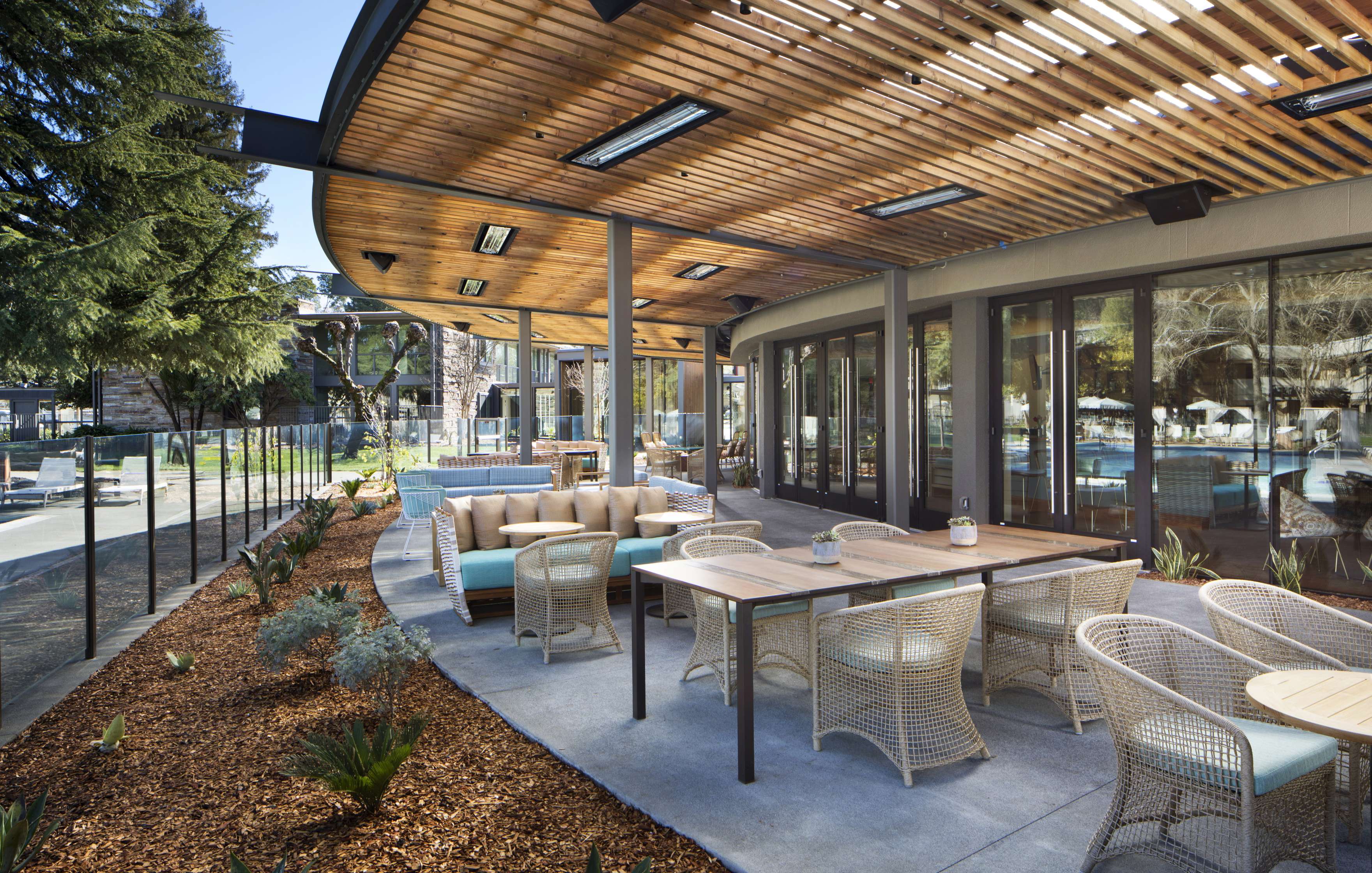 Outdoor patio seating area with modern wooden roof, a mix of tables and couches, and a view of a garden with a glass railing.