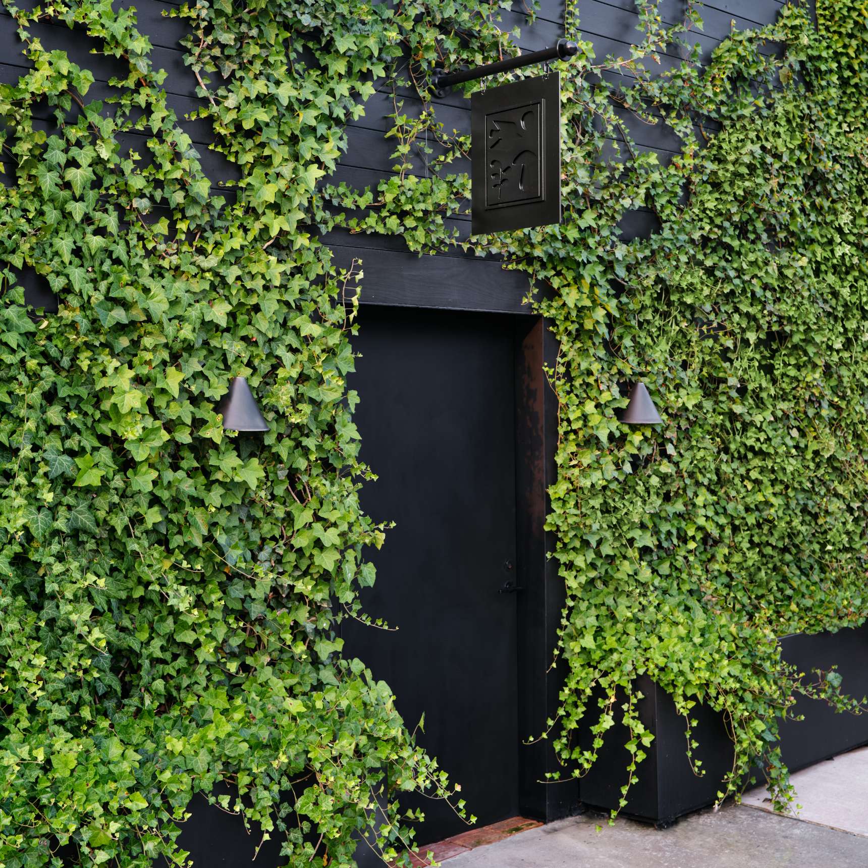 A black door is surrounded by a wall covered in green ivy. Two wall lamps frame the doorway, and a small black sign hangs above it.