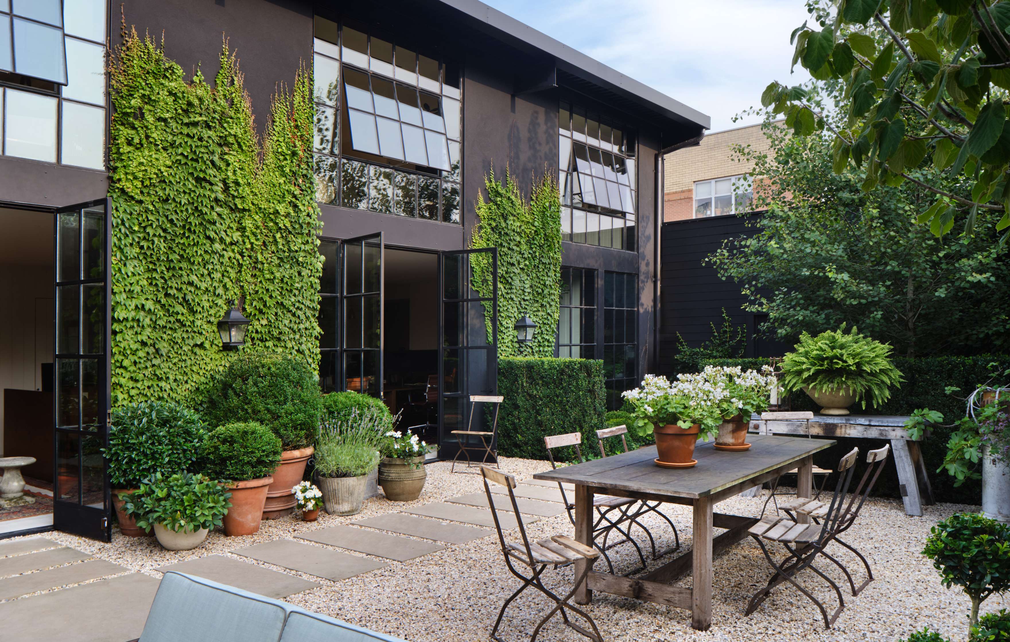 A modern courtyard with an outdoor seating area featuring a long table and chairs, surrounded by potted plants and ivy climbing a dark building with large windows.