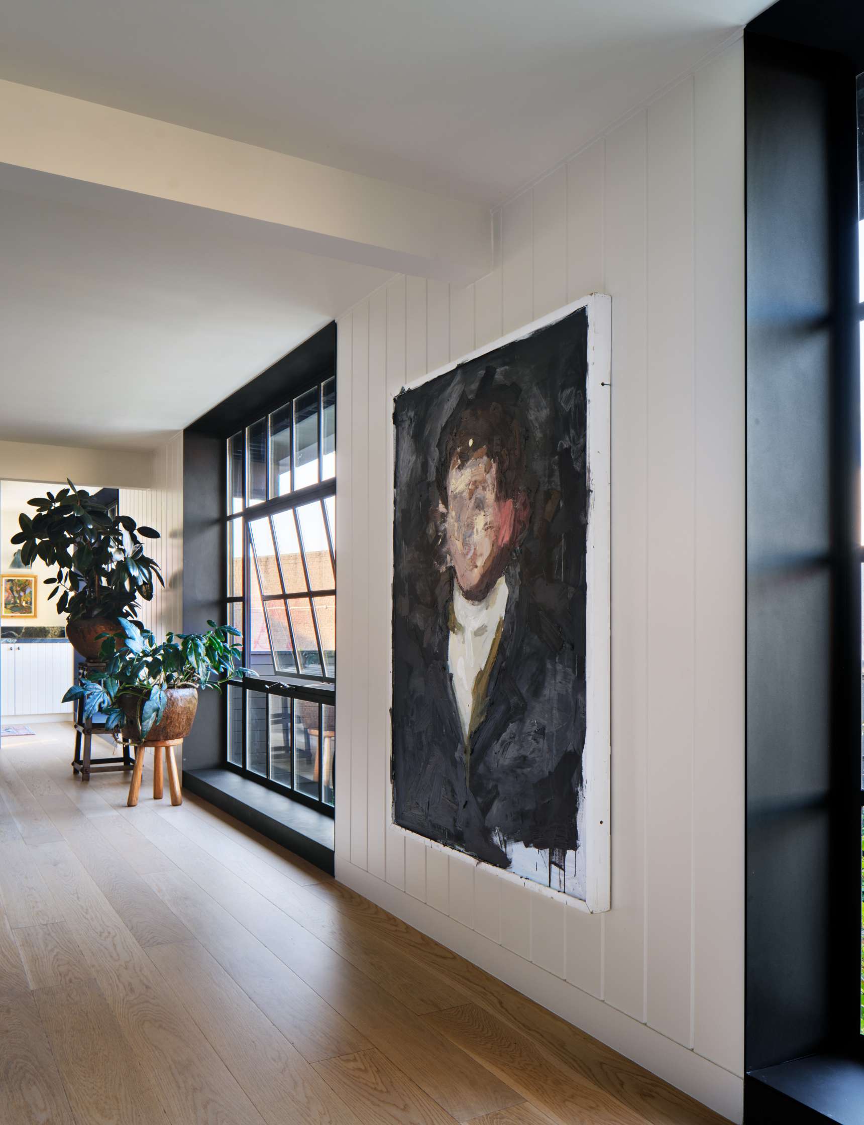 A dimly lit hallway features a large, abstract portrait hanging on a white wall beside a tall window. A potted plant sits on the floor near the window.