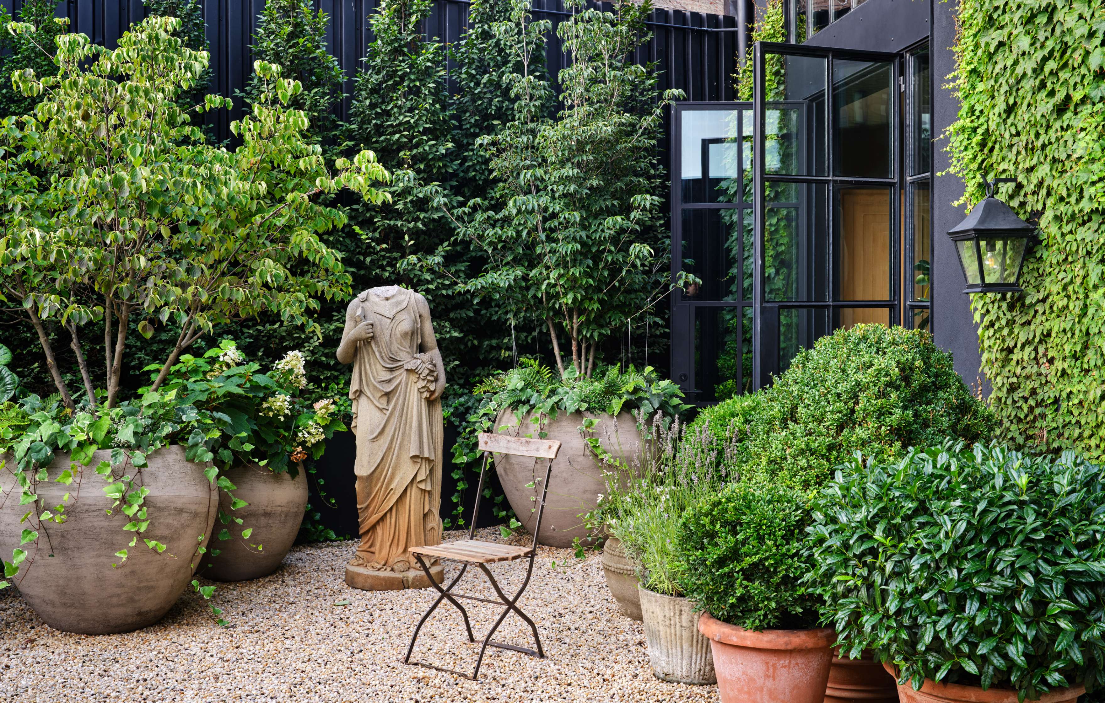 A garden with large potted plants, a headless statue, a wooden chair, and an open glass door on a building covered in ivy.