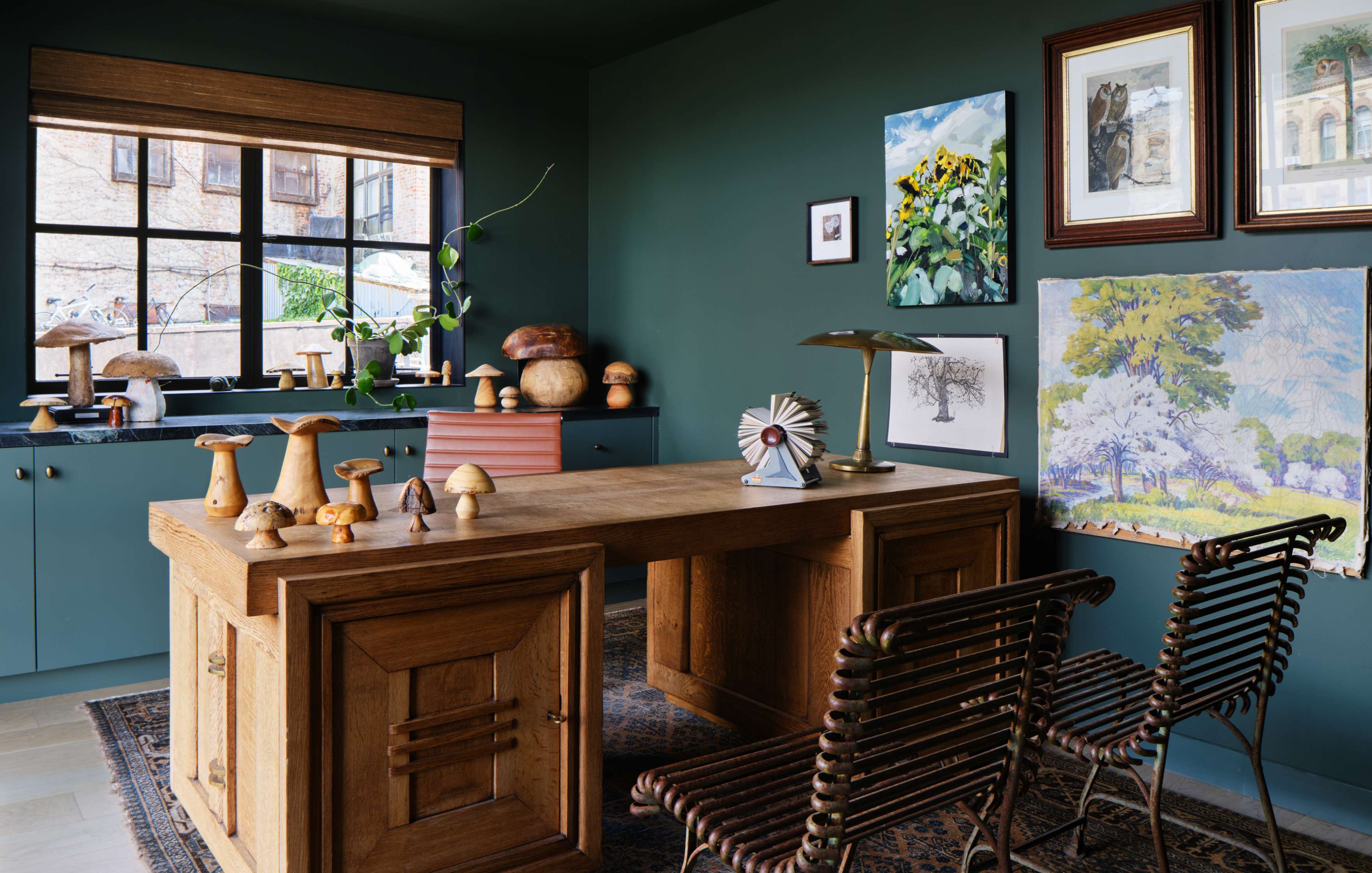 A home office with a wooden desk, two metal chairs, and artwork on dark green walls. The desk displays various decorative items, including mushroom figurines and a fan statue. A window with a bamboo shade overlooks outside.