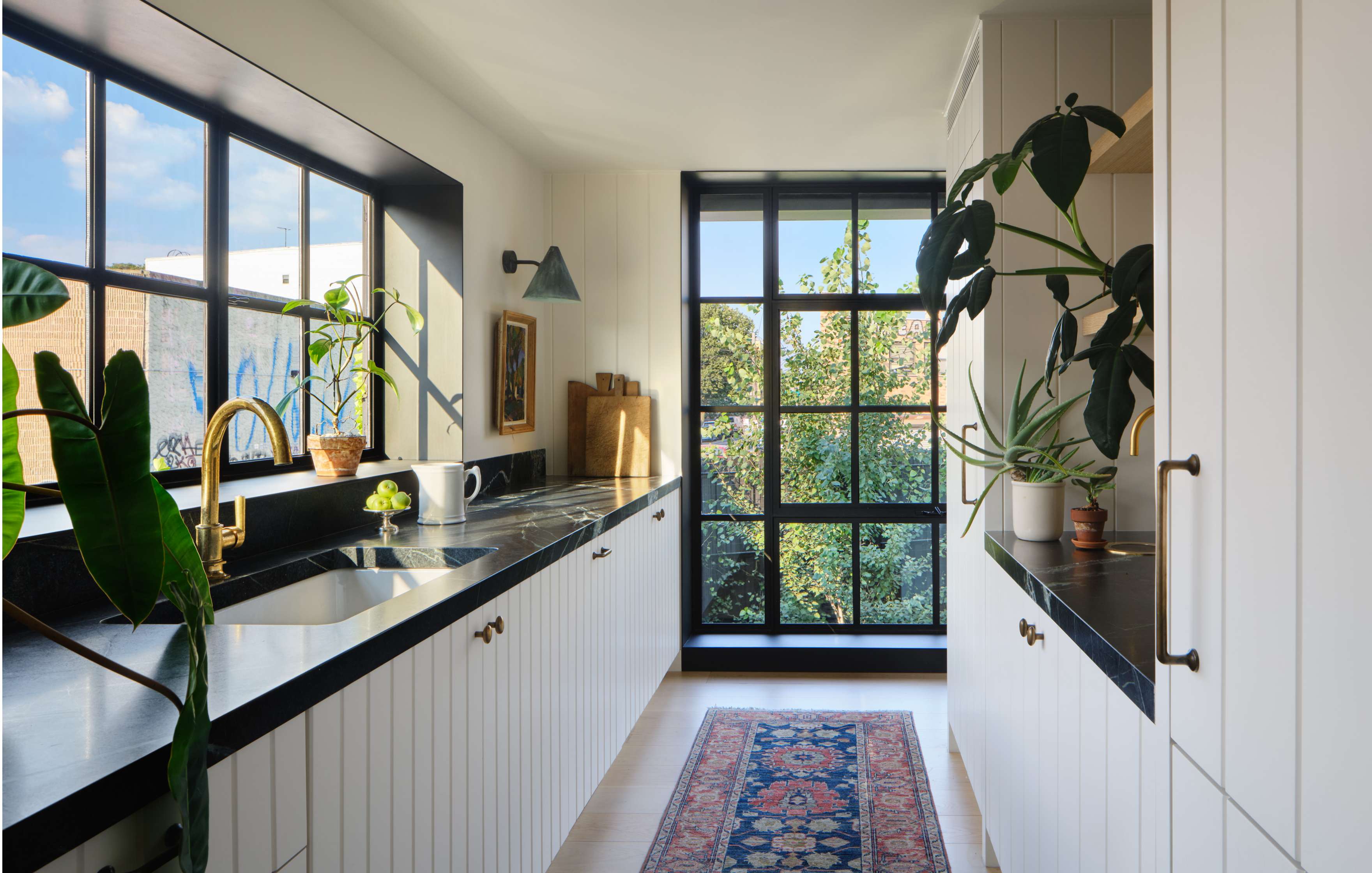 A modern kitchen with white cabinets, black countertops, large windows, leafy plants, and a colorful runner rug on the floor. Natural light is flooding through the windows.