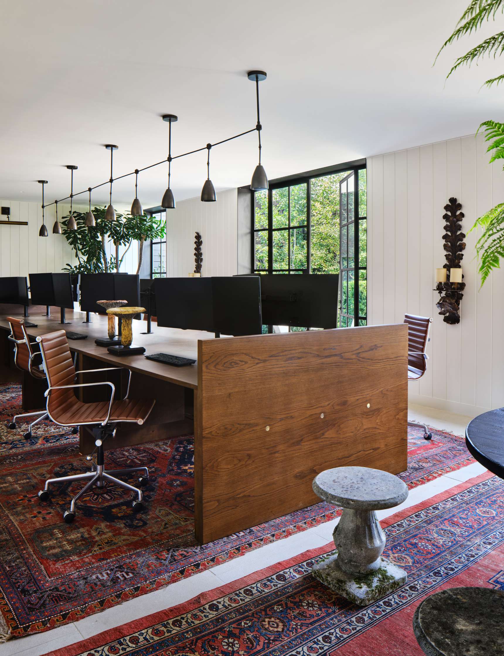 A modern office space with wooden desks, leather chairs, multiple computer monitors, pendant lighting, and a large window offering natural light, decorated with indoor plants and a patterned rug.