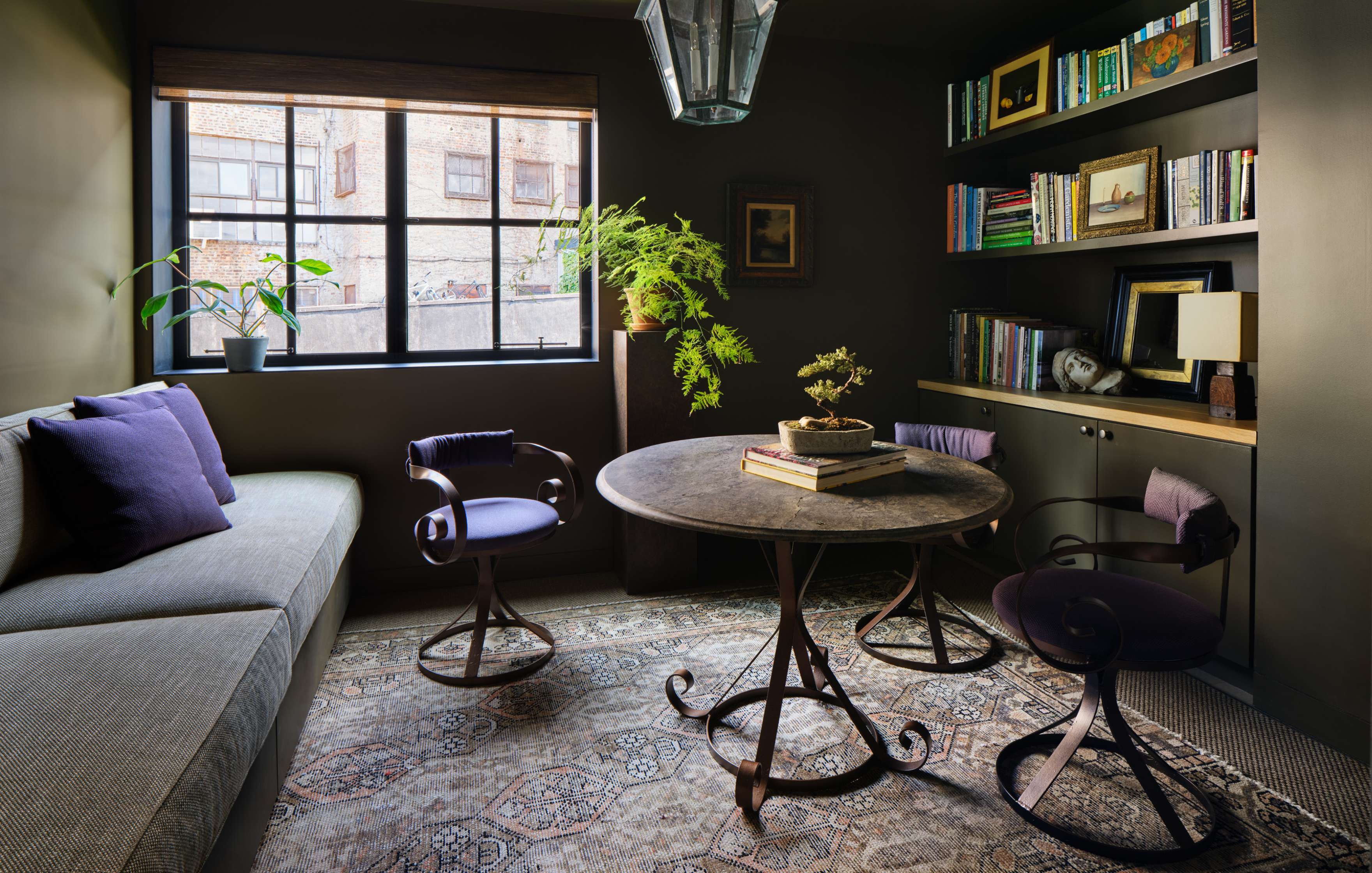 A cozy study room with a round table and three chairs, a couch, a large window, bookshelves, potted plants, and various books and decor items arranged neatly.