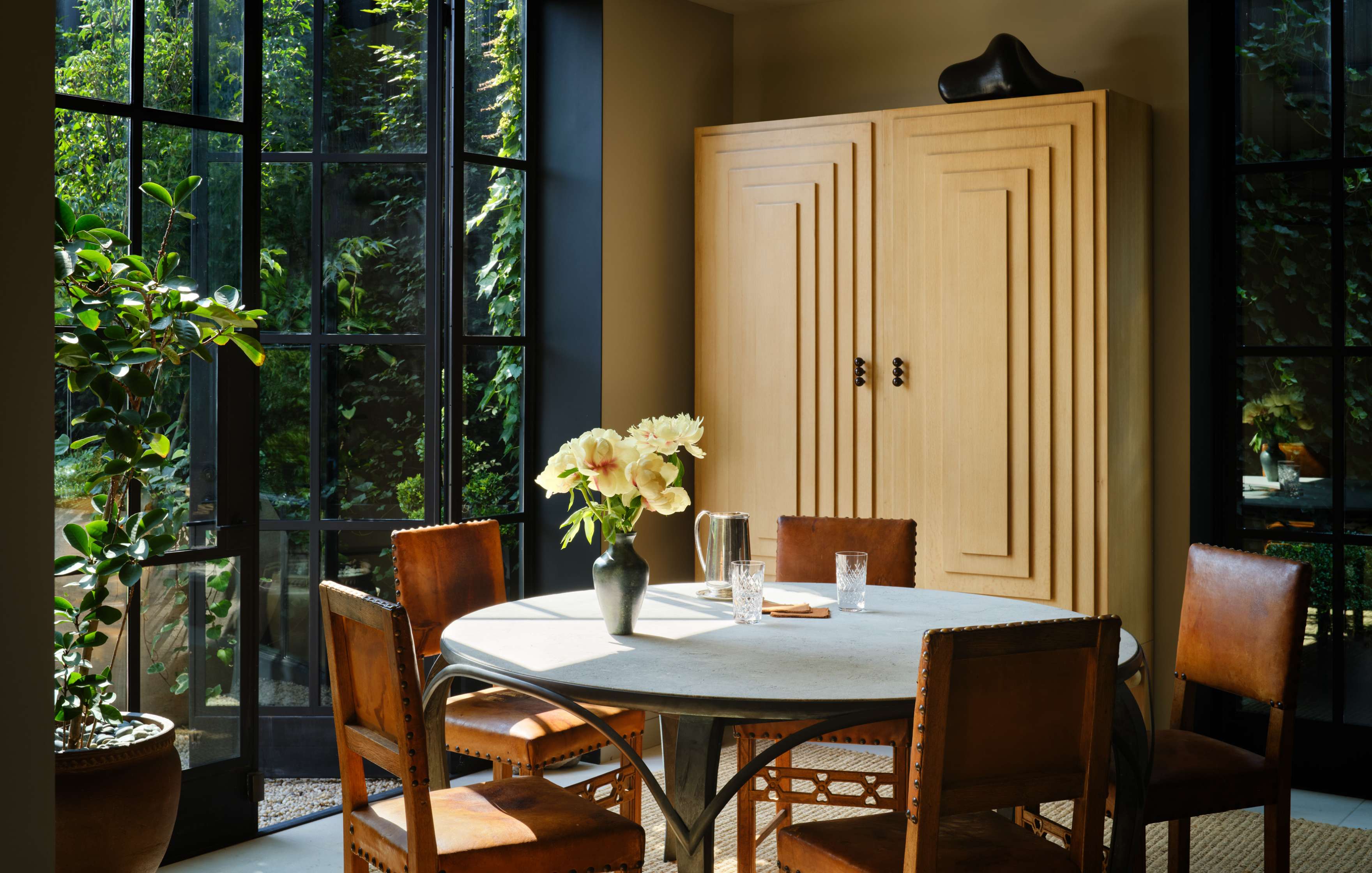 A cozy sunlit dining area with a round table, five chairs, a vase of flowers, and glassware. A wooden cabinet and large windows with garden views complete the setting.