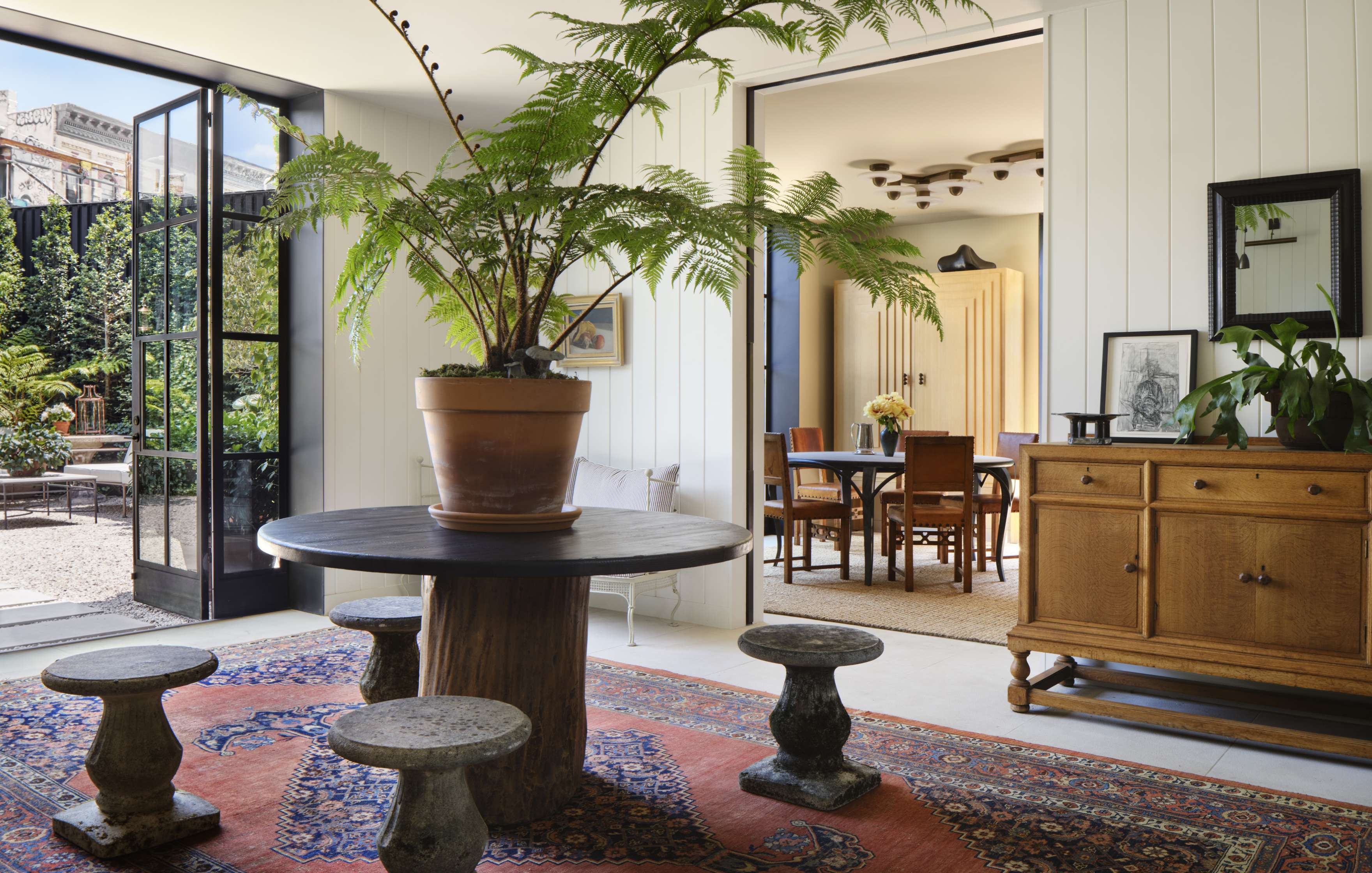 A sunlit room with a round table, potted fern, stone stools, oriental rug, and wooden sideboard. Adjacent room visible, furnished with a dining table and chairs, and large doors open to a garden.