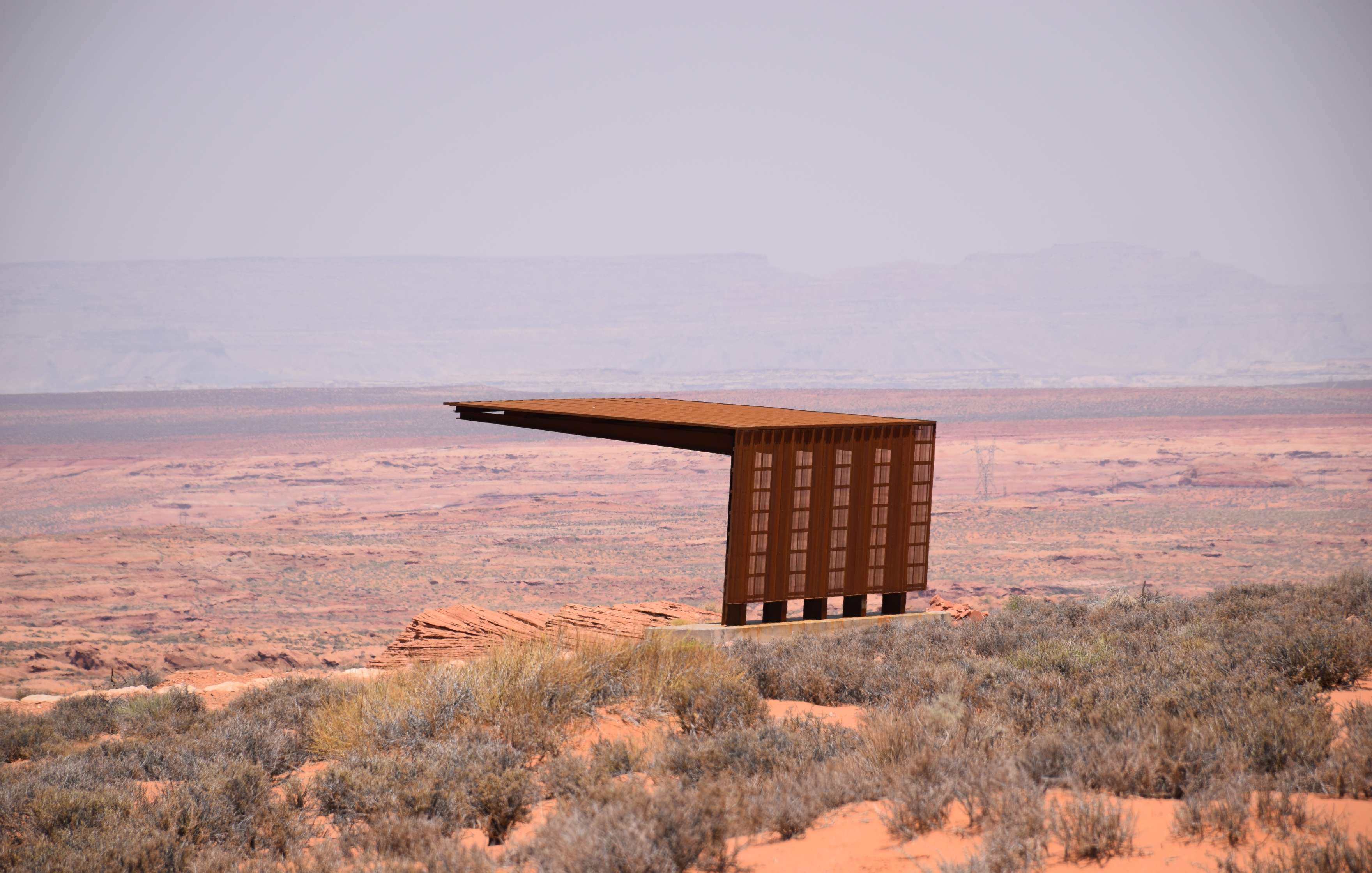 A modern, cantilevered viewing platform extends over a barren desert landscape under a hazy sky.