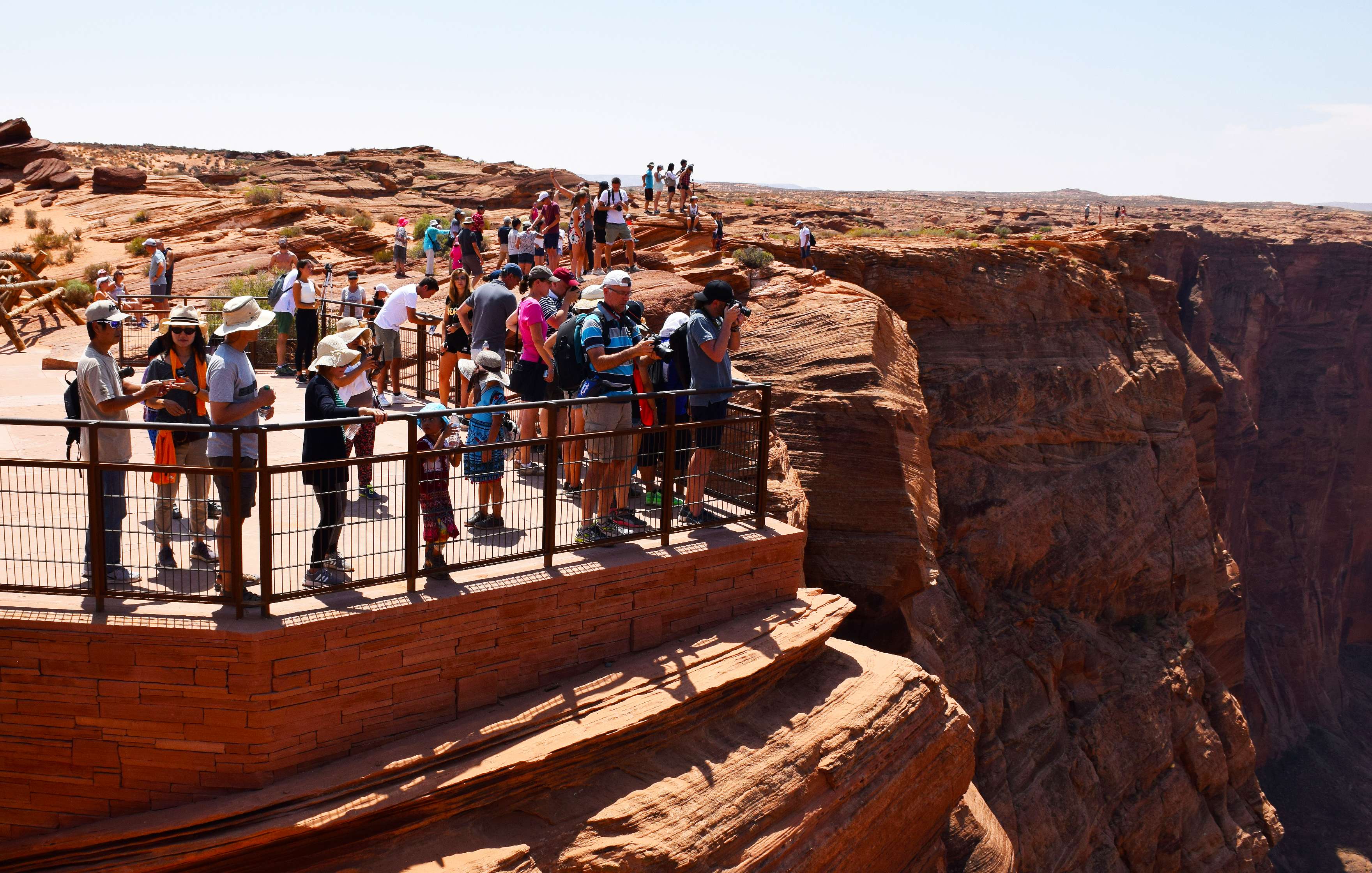 A group of people stand at a fenced viewpoint, overlooking the steep cliffs and expansive landscape of a canyon under a clear sky.