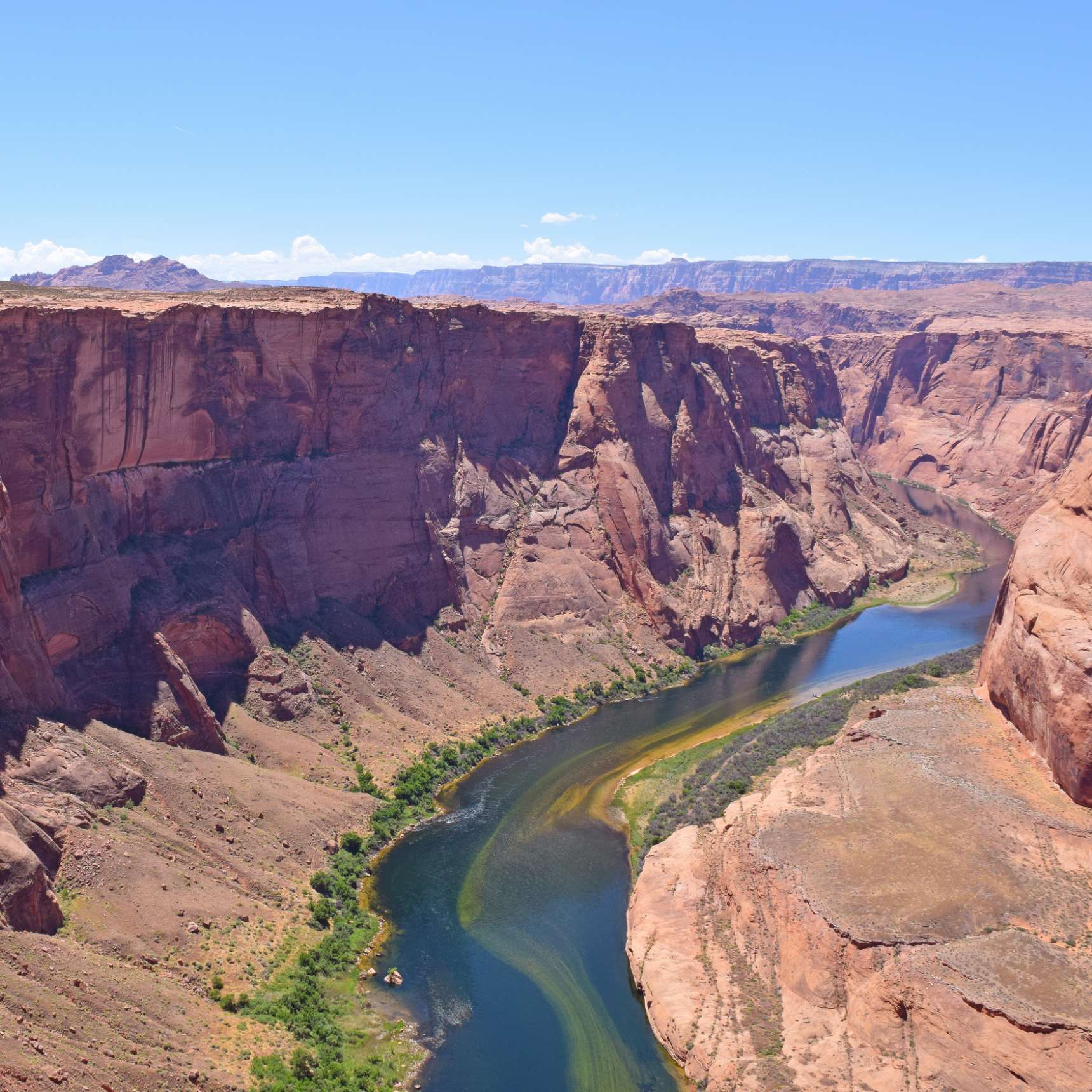 A winding river flows through a deep, rocky canyon with sheer red cliffs under a clear blue sky. Sparse vegetation lines the riverbanks.