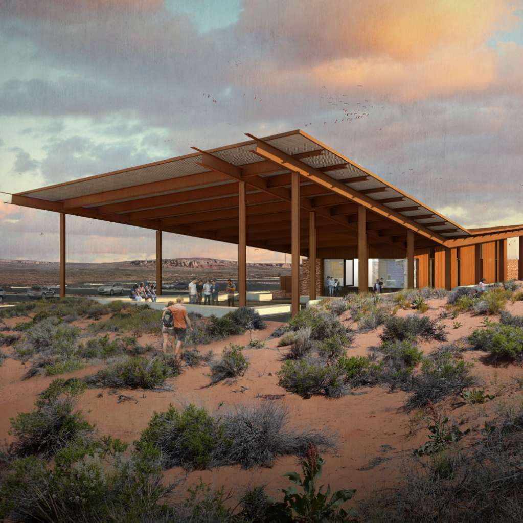 A modern structure with a broad, slatted roof stands in a desert landscape with shrubs, with people nearby and a colorful sky in the background.