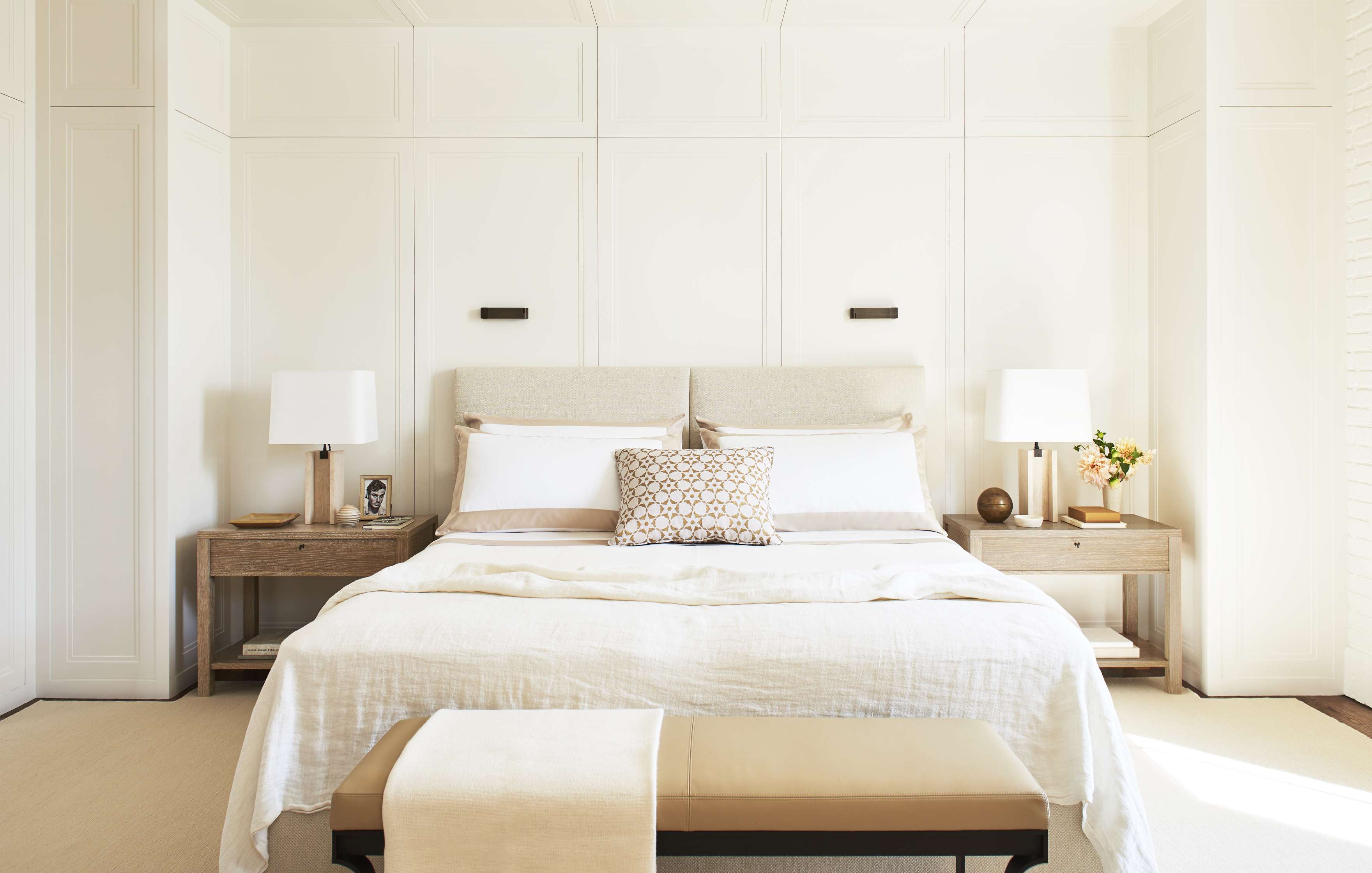 A neatly made bed with white linens and decorative pillows sits between two wooden nightstands. Each nightstand has a white lamp and other small items. A bench is positioned at the foot of the bed.