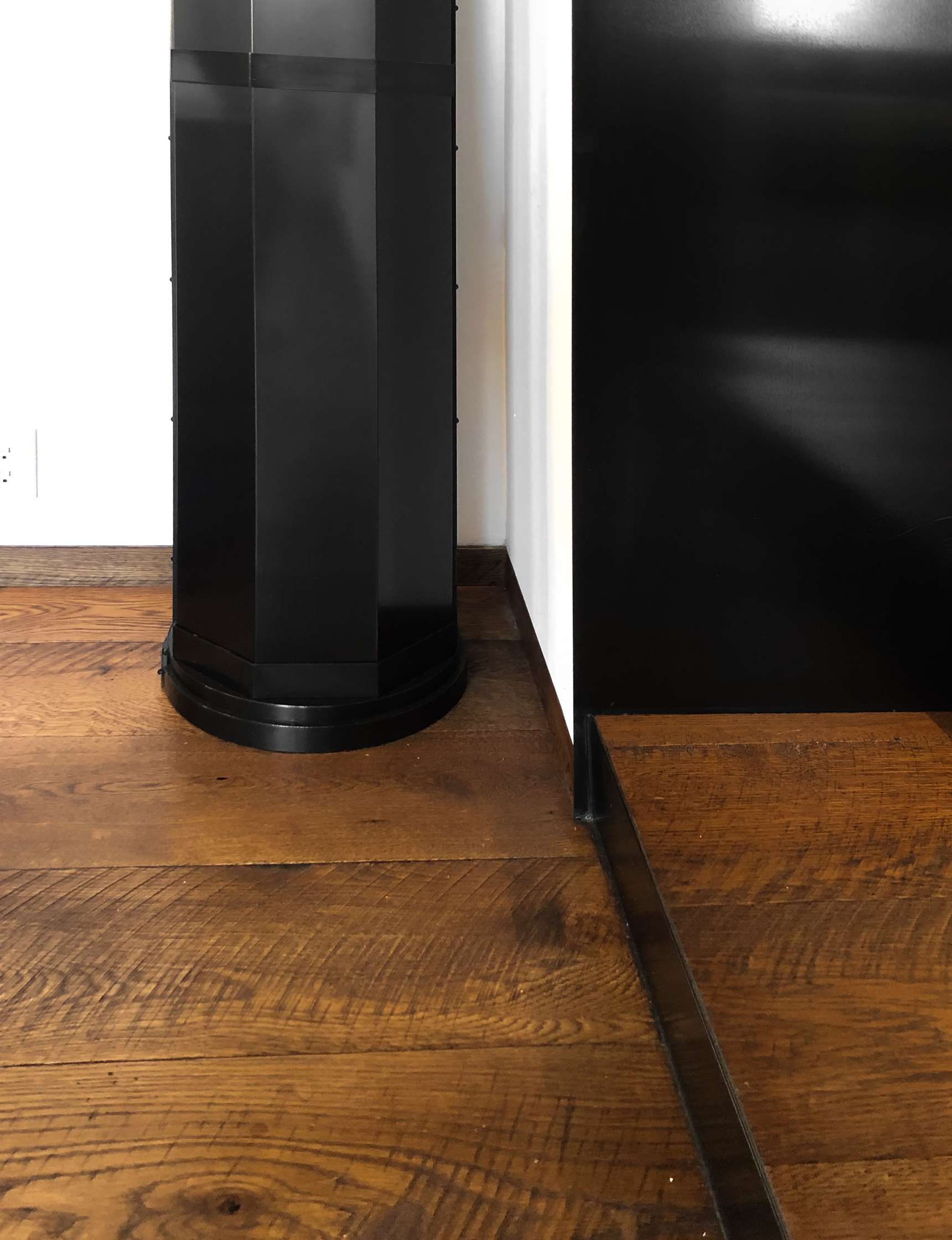 A black, vertical structure stands on a wooden floor next to a corner formed by white and black walls.