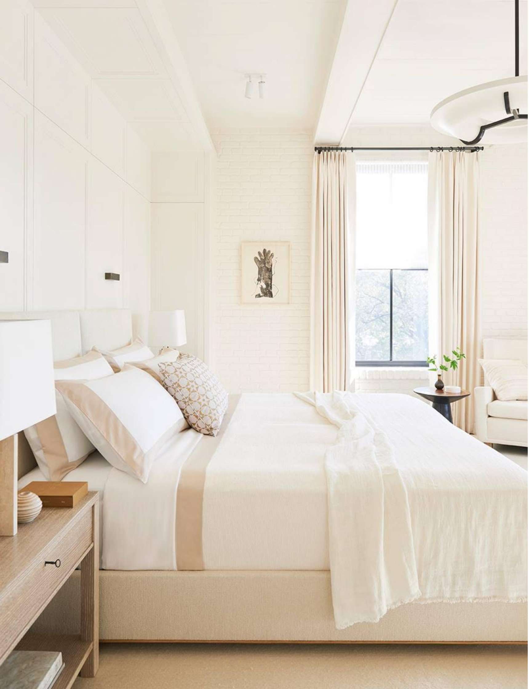 A bright bedroom with white walls, a large bed with white and beige bedding, a wooden bedside table with a lamp, and a window framed by light curtains.