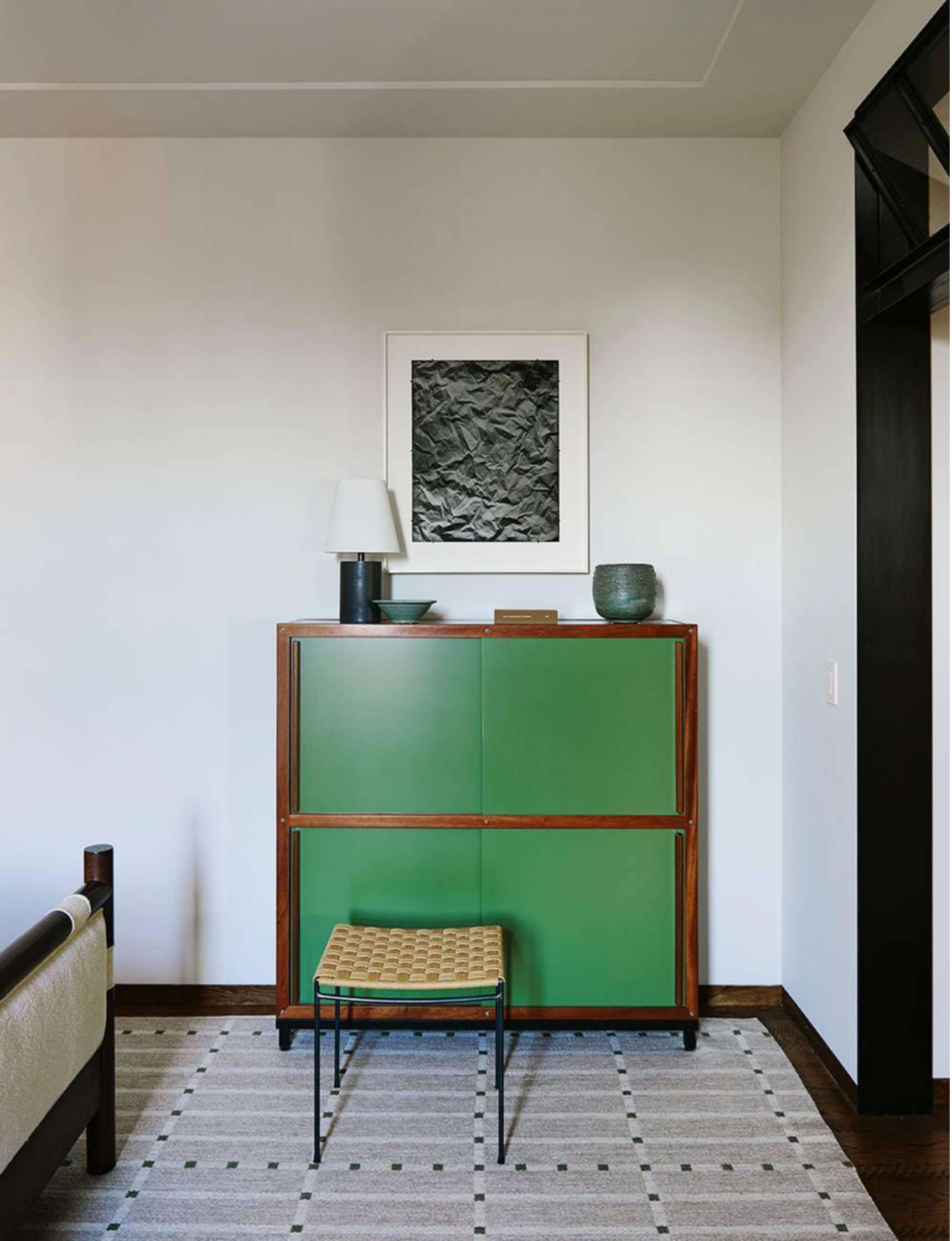 A room corner featuring a green cabinet with wooden trim, a lamp, decorative vases on top, and a framed abstract artwork above. A woven stool sits in front of the cabinet on a patterned rug.