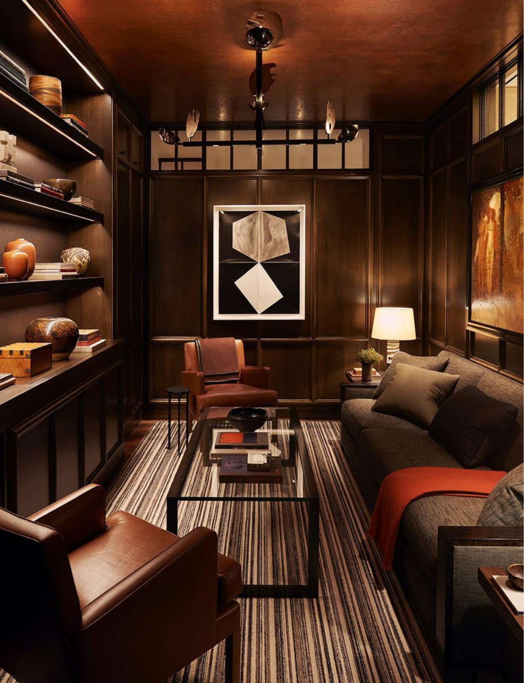 A cozy room with dark wood-paneled walls, a striped rug, a glass coffee table, leather chairs, a sofa, shelves with decor, and framed art on the walls. There is a lit table lamp on the right.