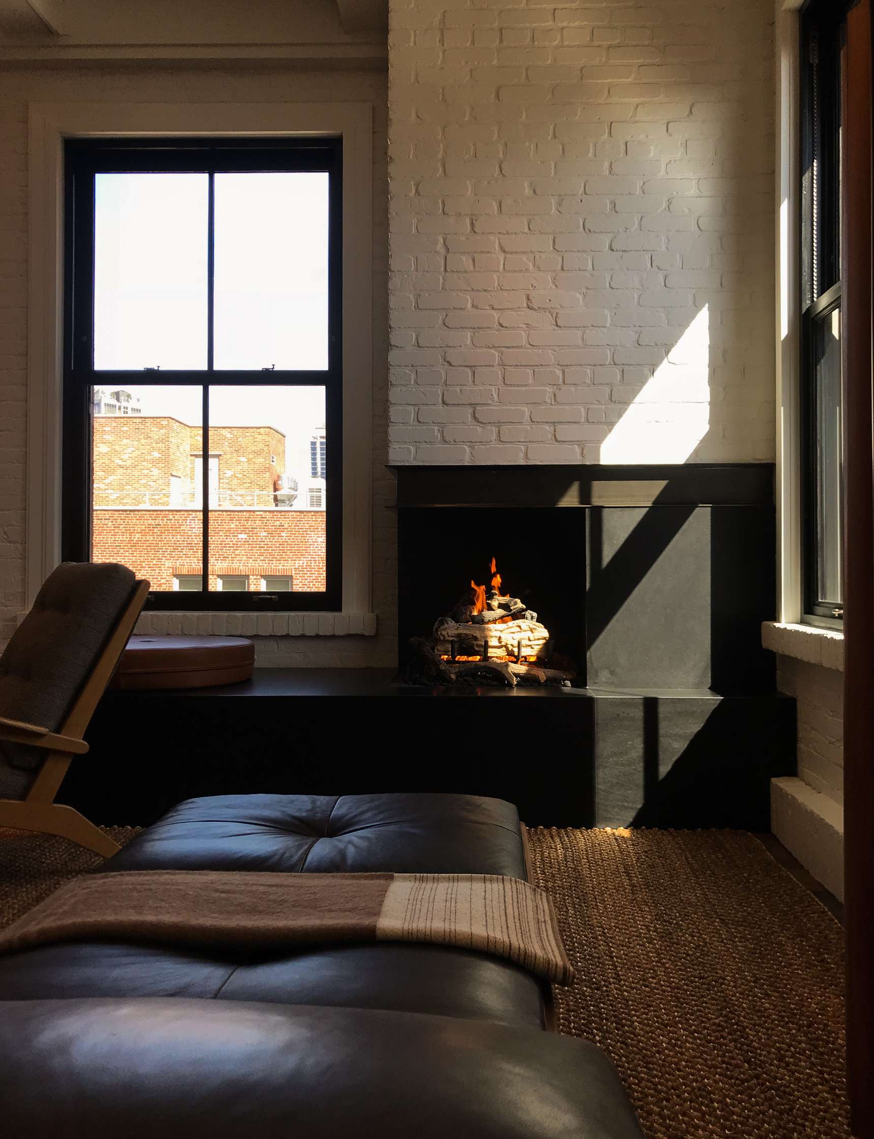 A cozy room featuring a leather sofa, a window allowing sunlight to stream in, and a fireplace with a burning fire. The exposed brick walls add a rustic touch.