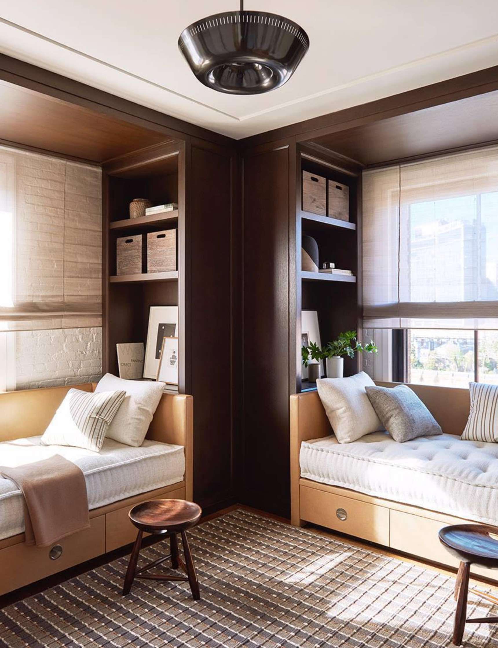 A cozy room with two built-in wooden daybeds, storage shelves above, and a round ceiling light. The beds are adorned with cushions, and a striped rug is placed between them. Large windows let in ample light.