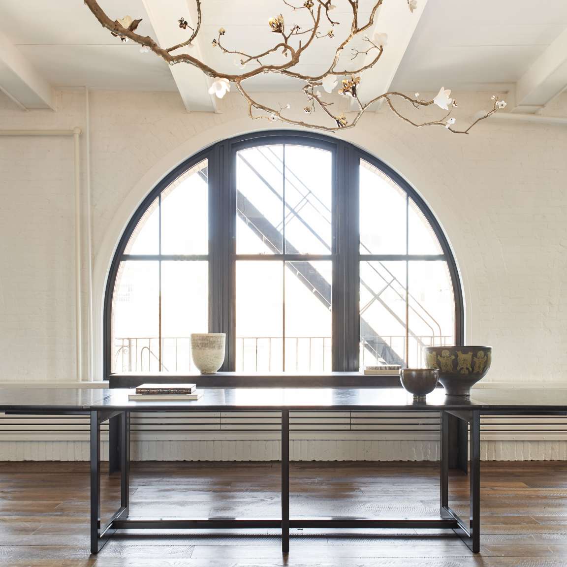 A minimalist interior with a large arched window, a black table, and a few decorative items. A ceiling light fixture has branches with white flowers. The floor is wooden.