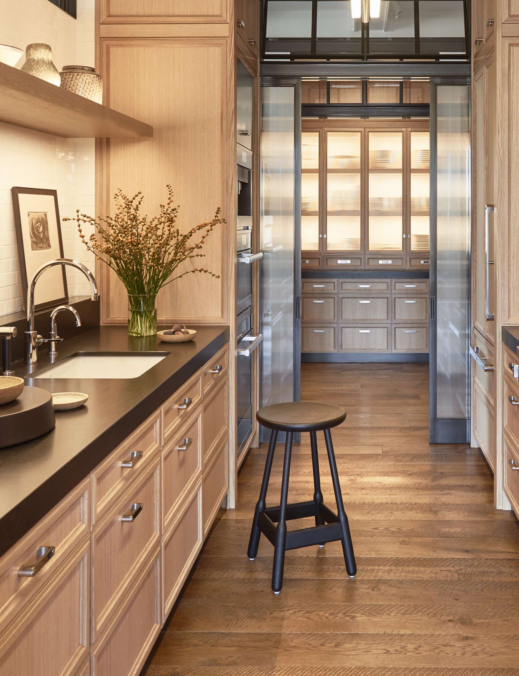 A modern kitchen with wooden cabinets, a black countertop with a sink, a central black stool, open shelves with baskets, and a corridor leading to a pantry with lit glass cabinets.