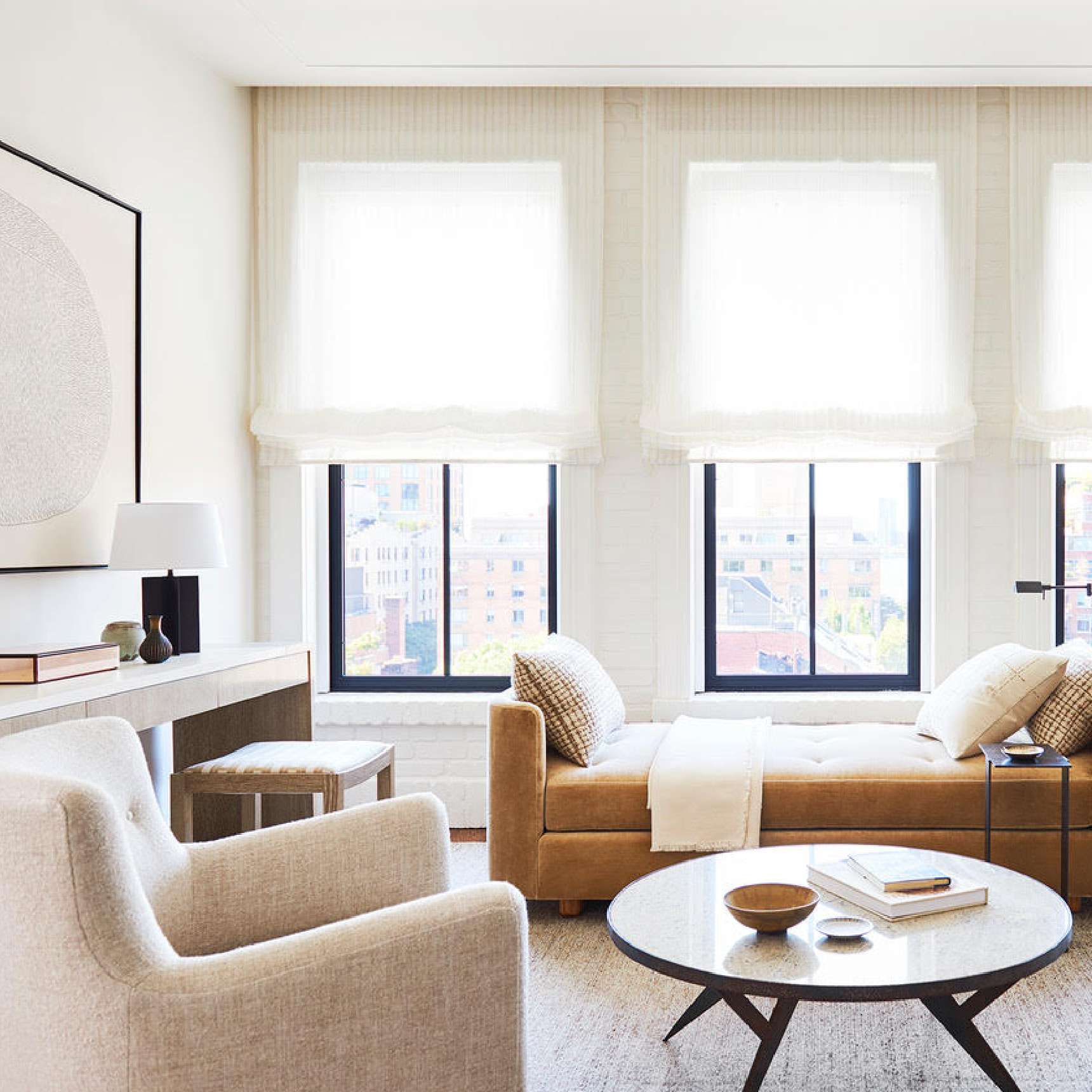 A modern living room with large windows, white walls, a beige armchair, a wooden coffee table with books, and a beige chaise lounge. Natural light fills the room.