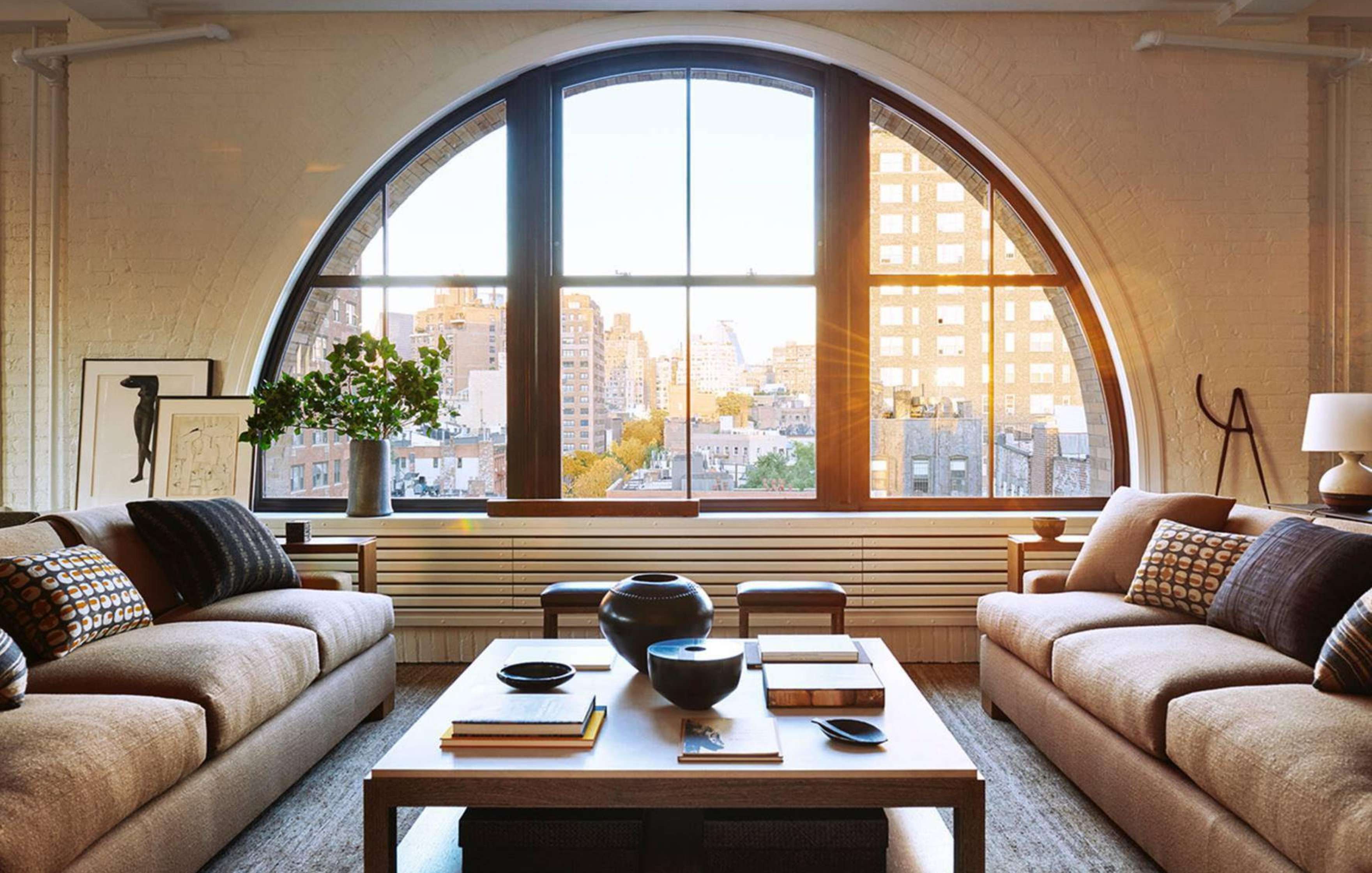 A modern living room with two beige couches, a wooden coffee table, and a large arched window offering a cityscape view. Decor includes plants, artwork, and various tabletop items.