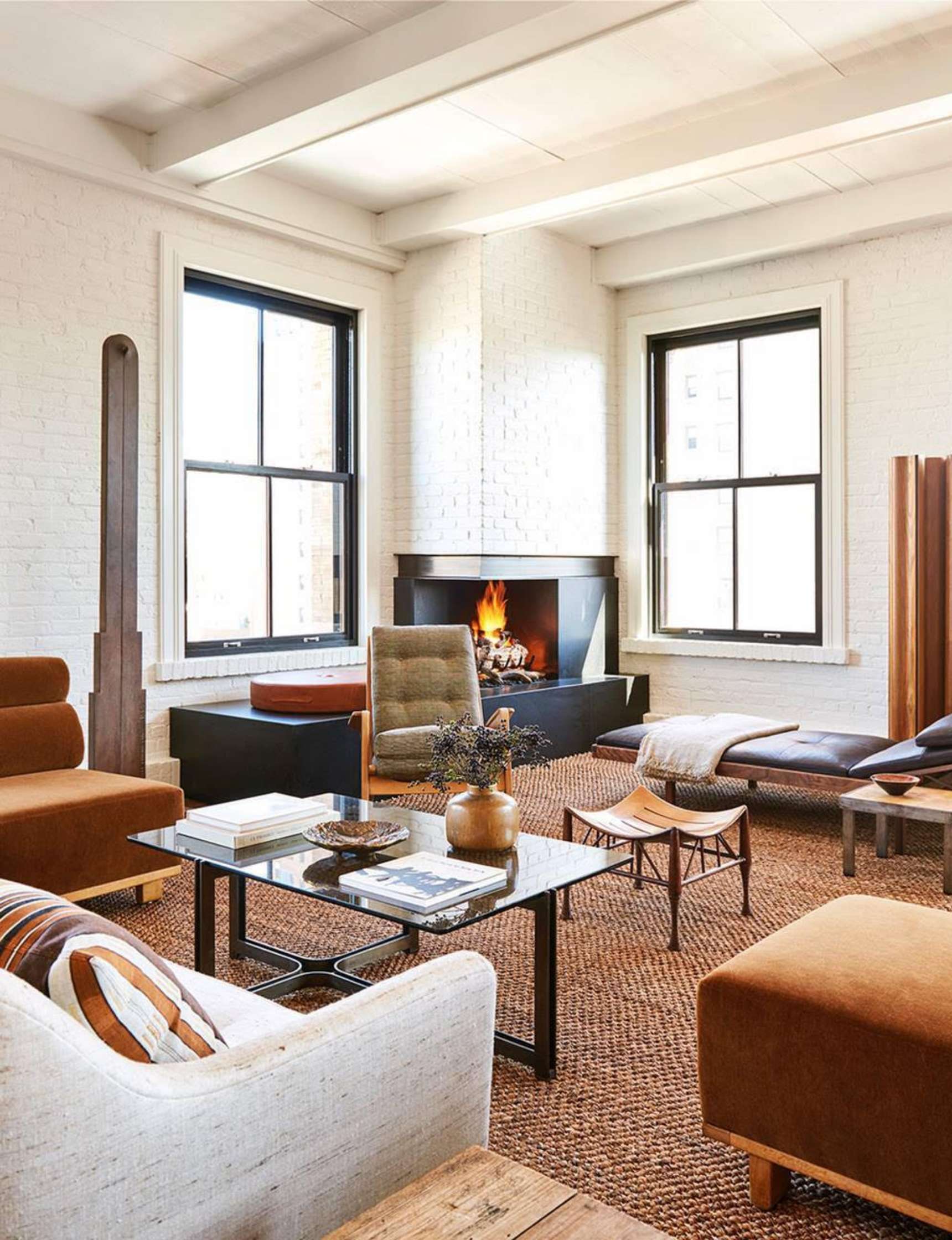 A modern living room features a fireplace, two windows, assorted brown and beige furniture, a glass coffee table, and decorative items on a woven rug.