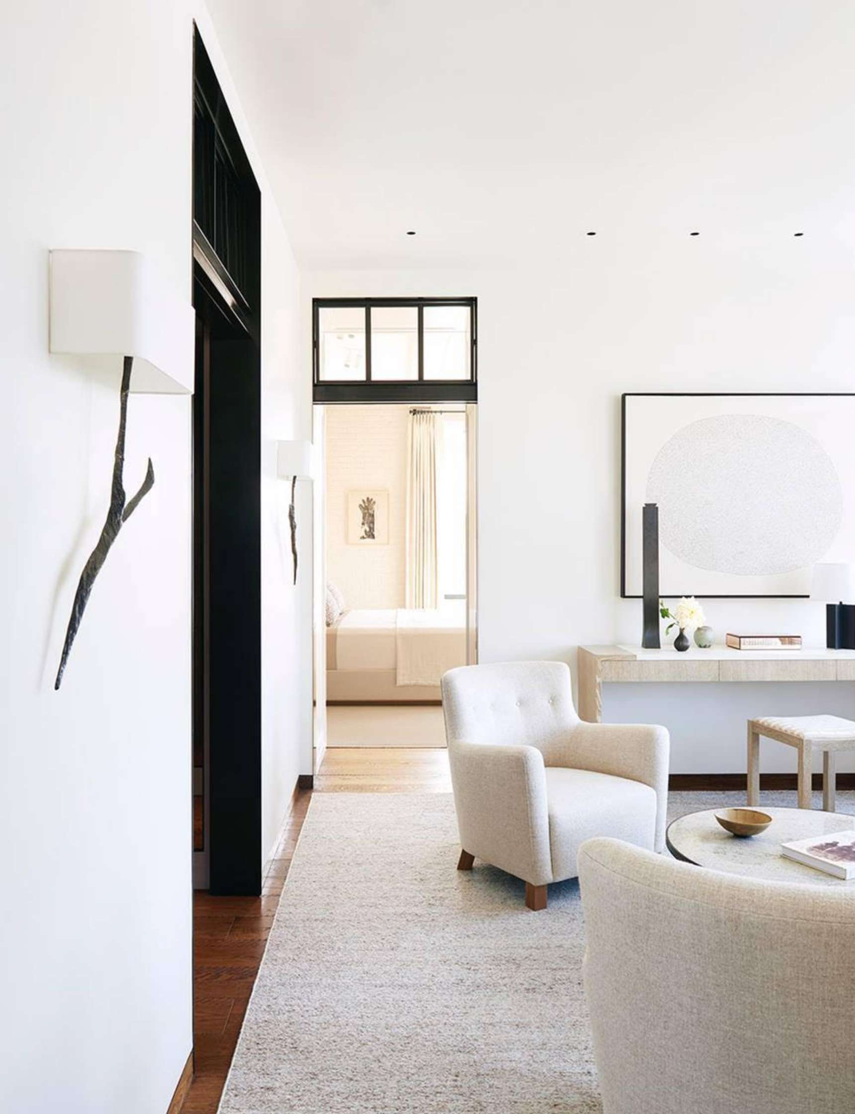 A modern living room with white walls, light furniture, a round coffee table, and a large abstract wall art piece. A doorway leads to a bright bedroom with a bed and a window with light curtains.