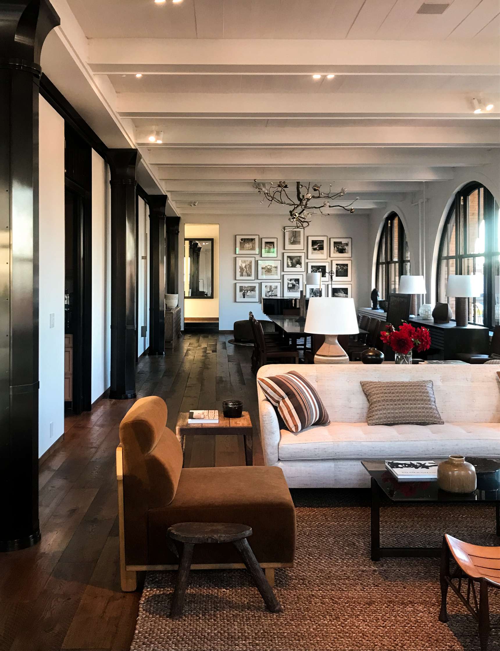 A modern living room with white walls, exposed beams, and dark wood flooring. It features a beige sofa, brown chair, small table, and framed pictures on the wall. Large windows allow natural light in.