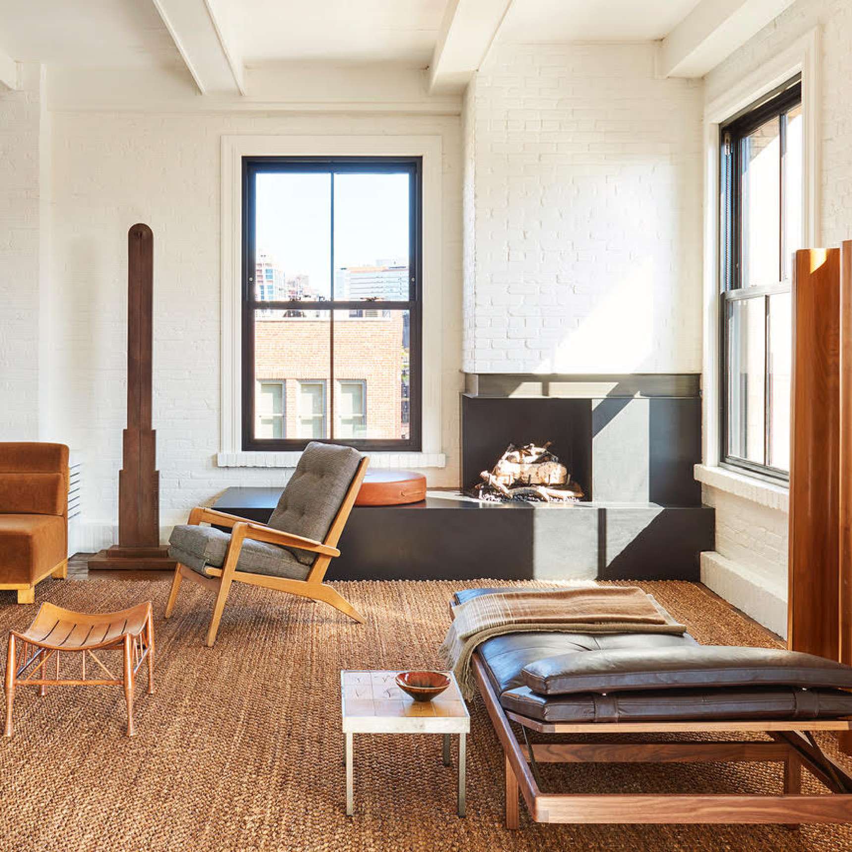 A minimalist living room with a fireplace, wooden furniture, a jute rug, and a large window letting in natural light.