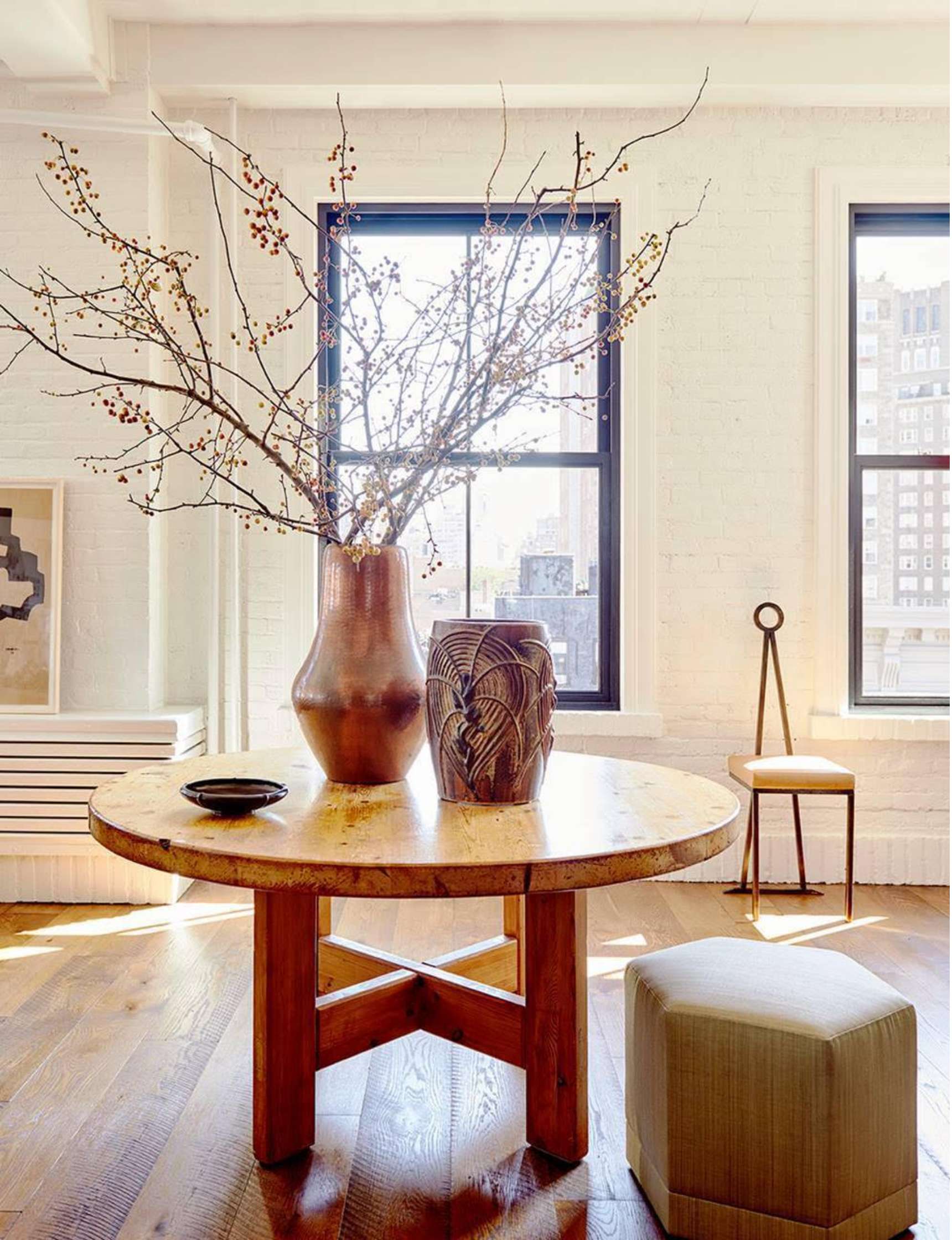 A round wooden table with vases and branches sits in a bright room with two large windows, a chair, and a small ottoman.