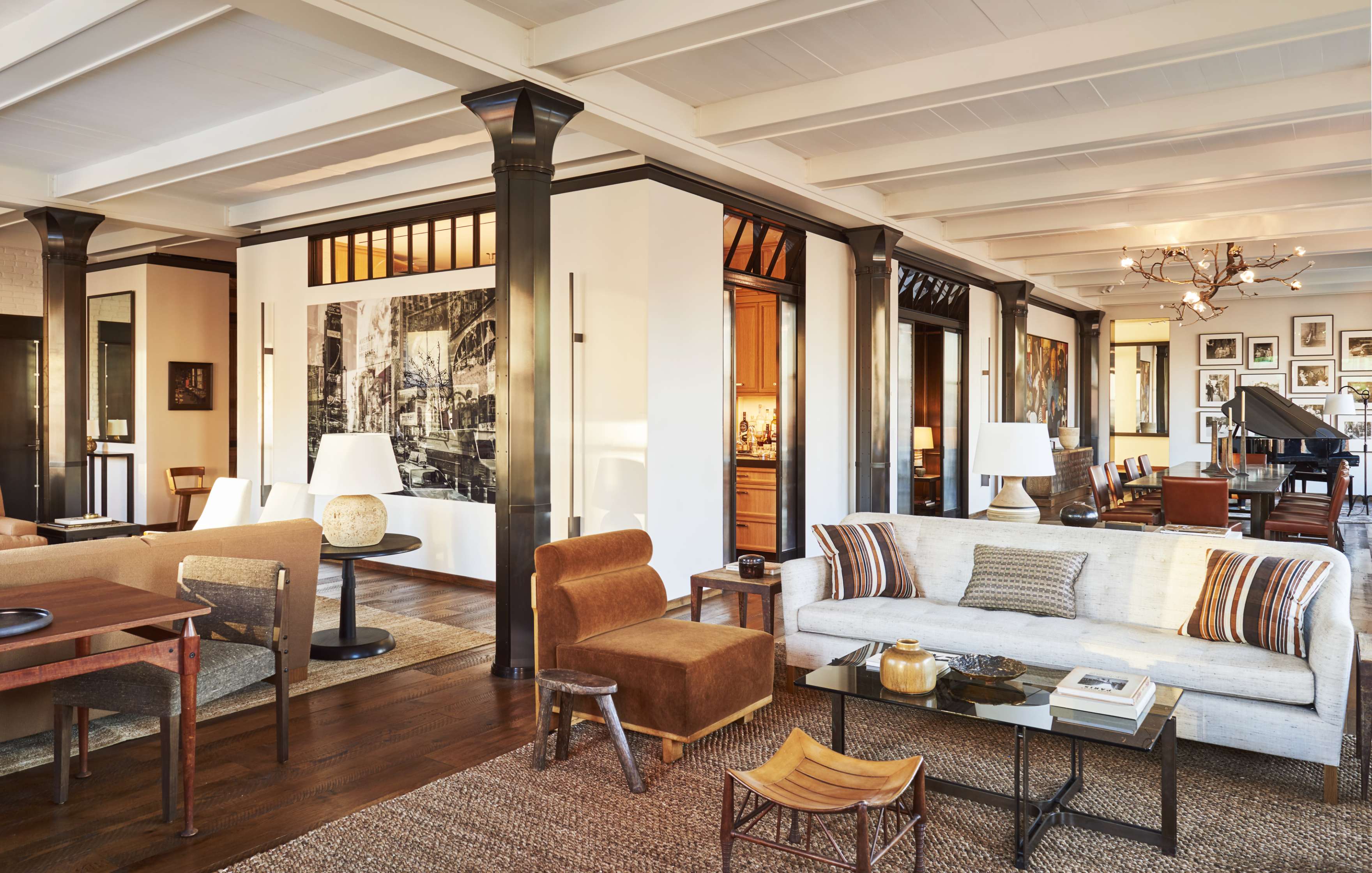 A spacious, well-lit living room with a white sofa, brown chairs, a coffee table, and modern decor. A dining area and a piano are visible in the background, along with various framed pictures on the walls.