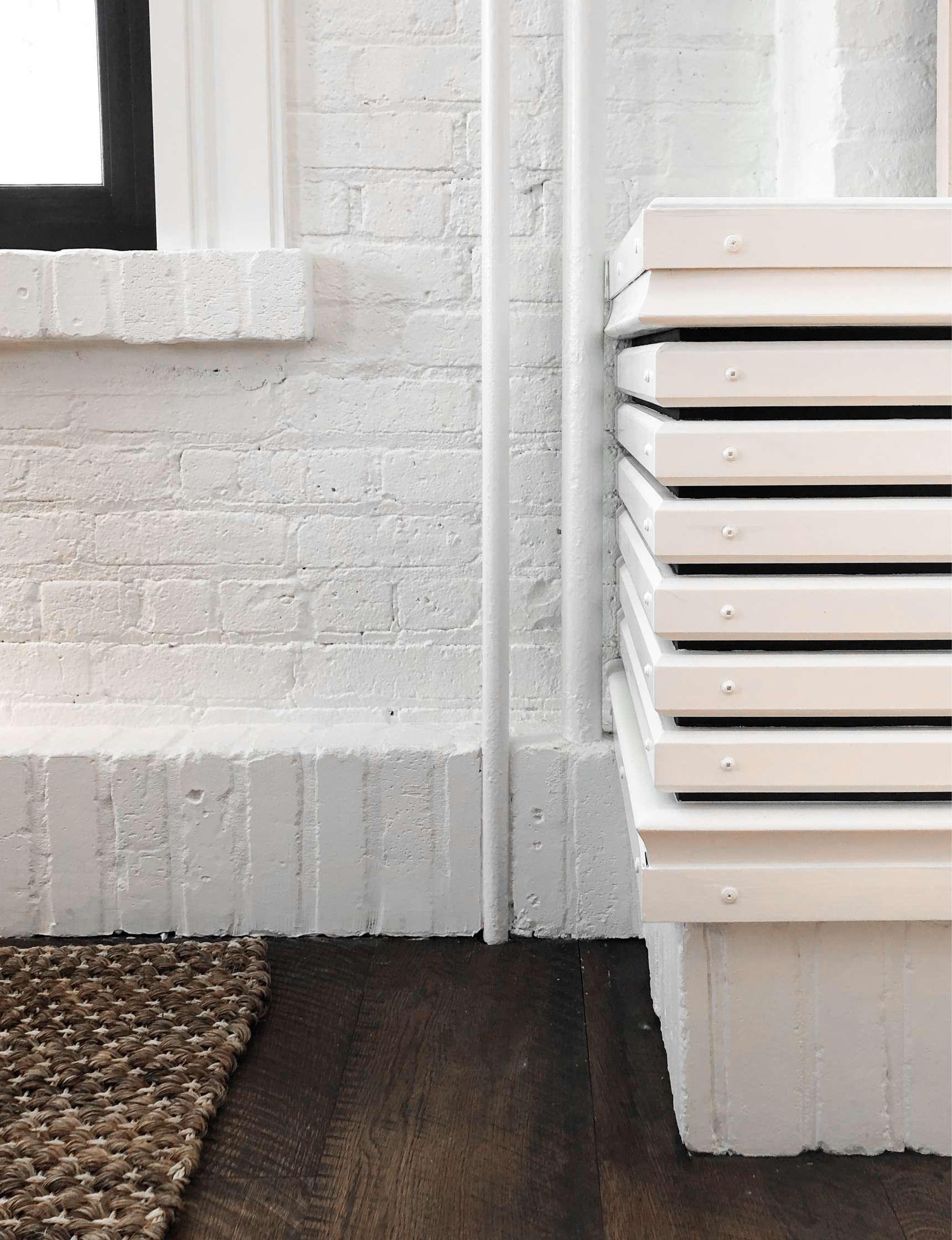 A white radiator is mounted on a white brick wall beside a black-framed window, with a textured beige rug on a dark wooden floor.