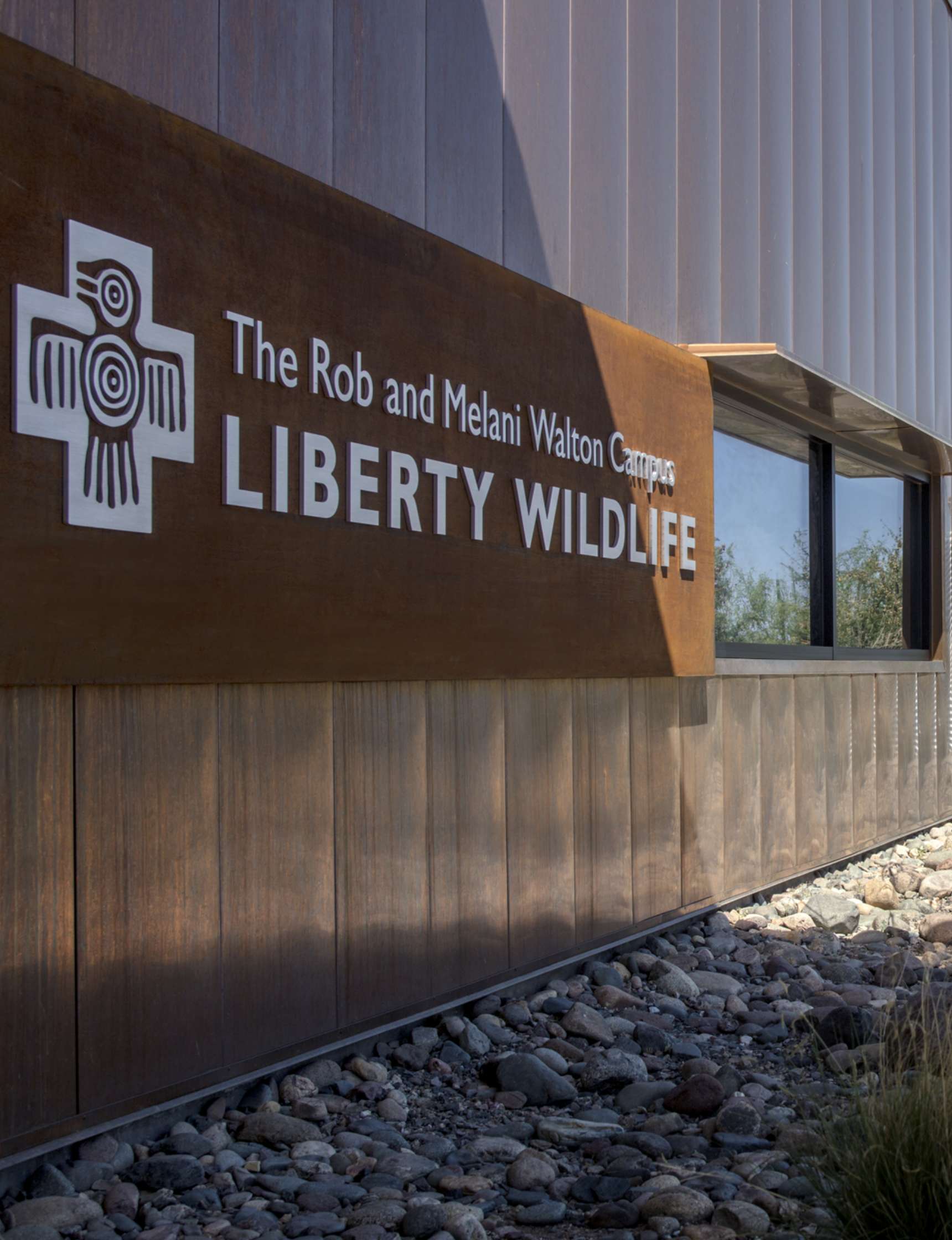 Sign for The Rob and Melani Walton Campus, Liberty Wildlife, mounted on an exterior wall with windows and a gravel area below.