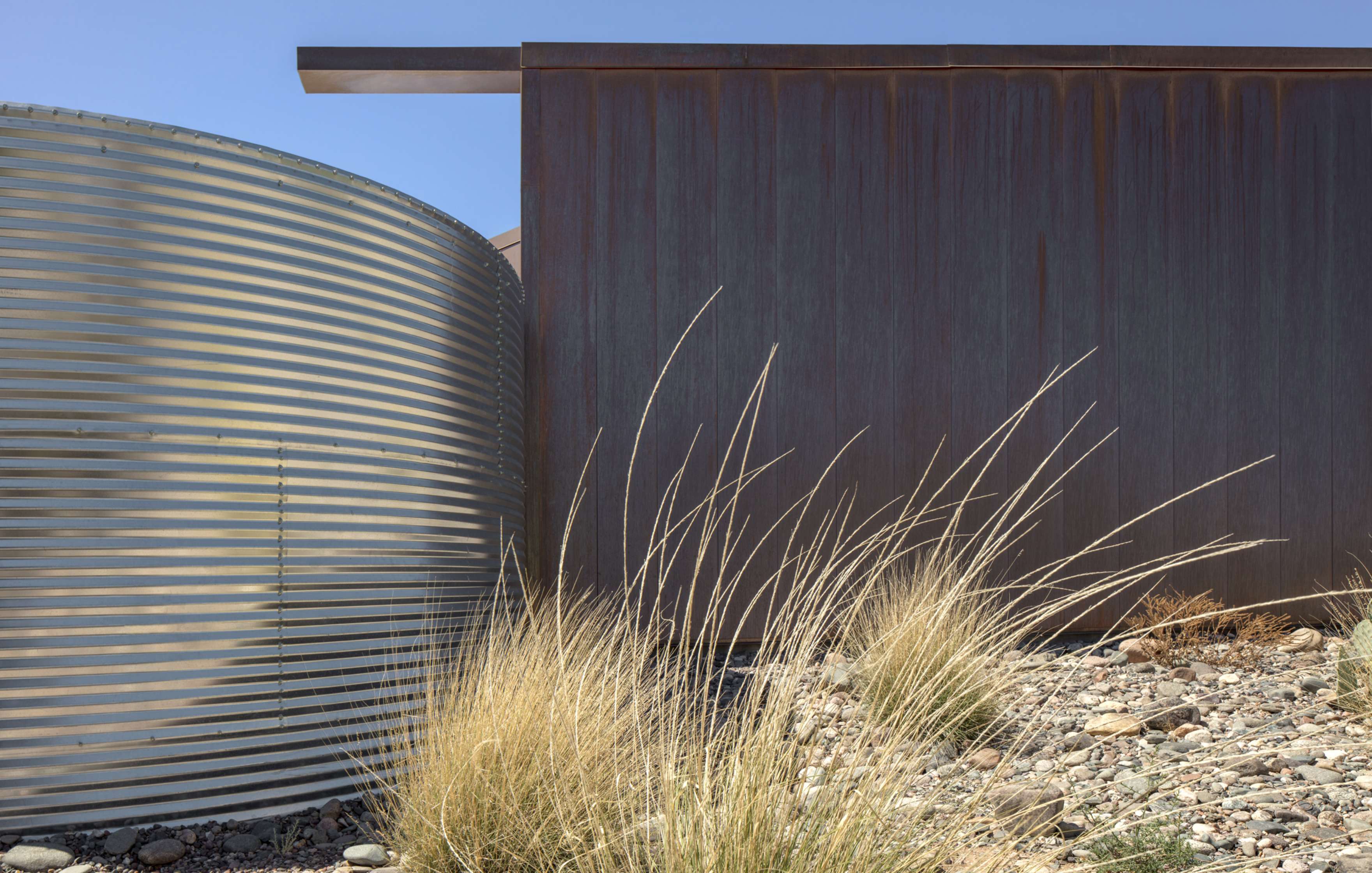 A modern metal building with a corrugated structure and a rusted wall stands next to dry grasses and rocky ground, against a clear blue sky.