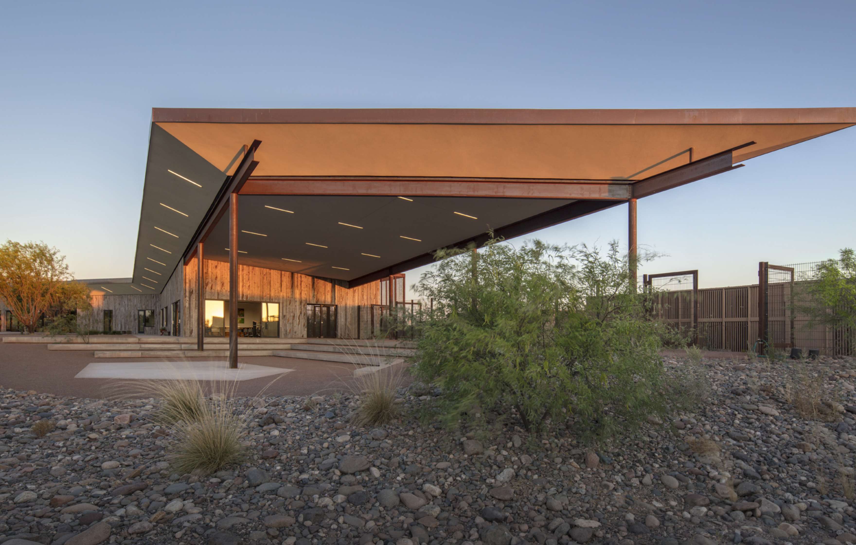 Modern building with a large overhanging roof, surrounded by desert landscape with rocks and shrubs, under a clear sky at sunset.