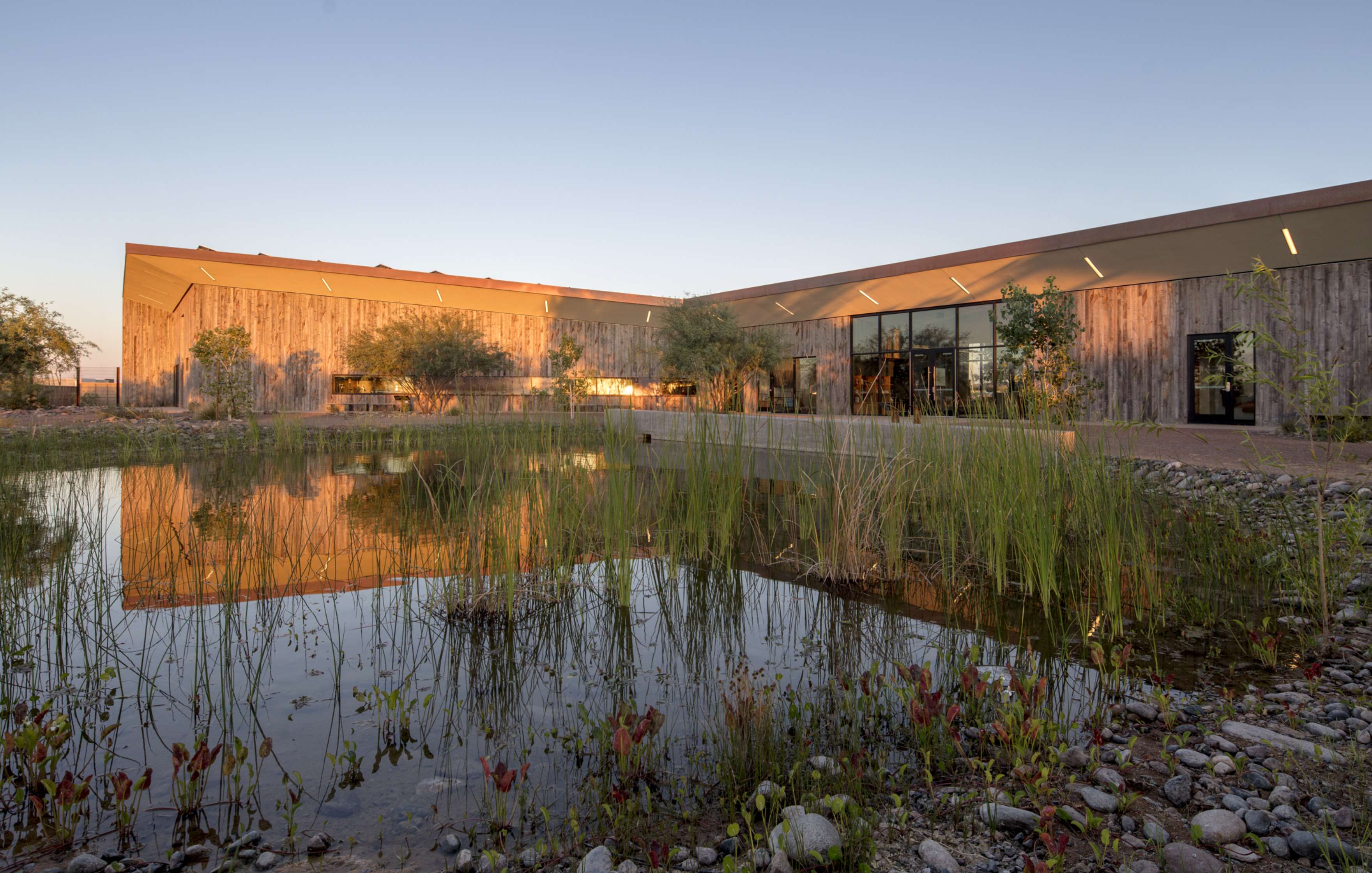 A modern building with large glass windows and wooden exterior is reflected in a pond, surrounded by sparse vegetation and trees under a clear sky.