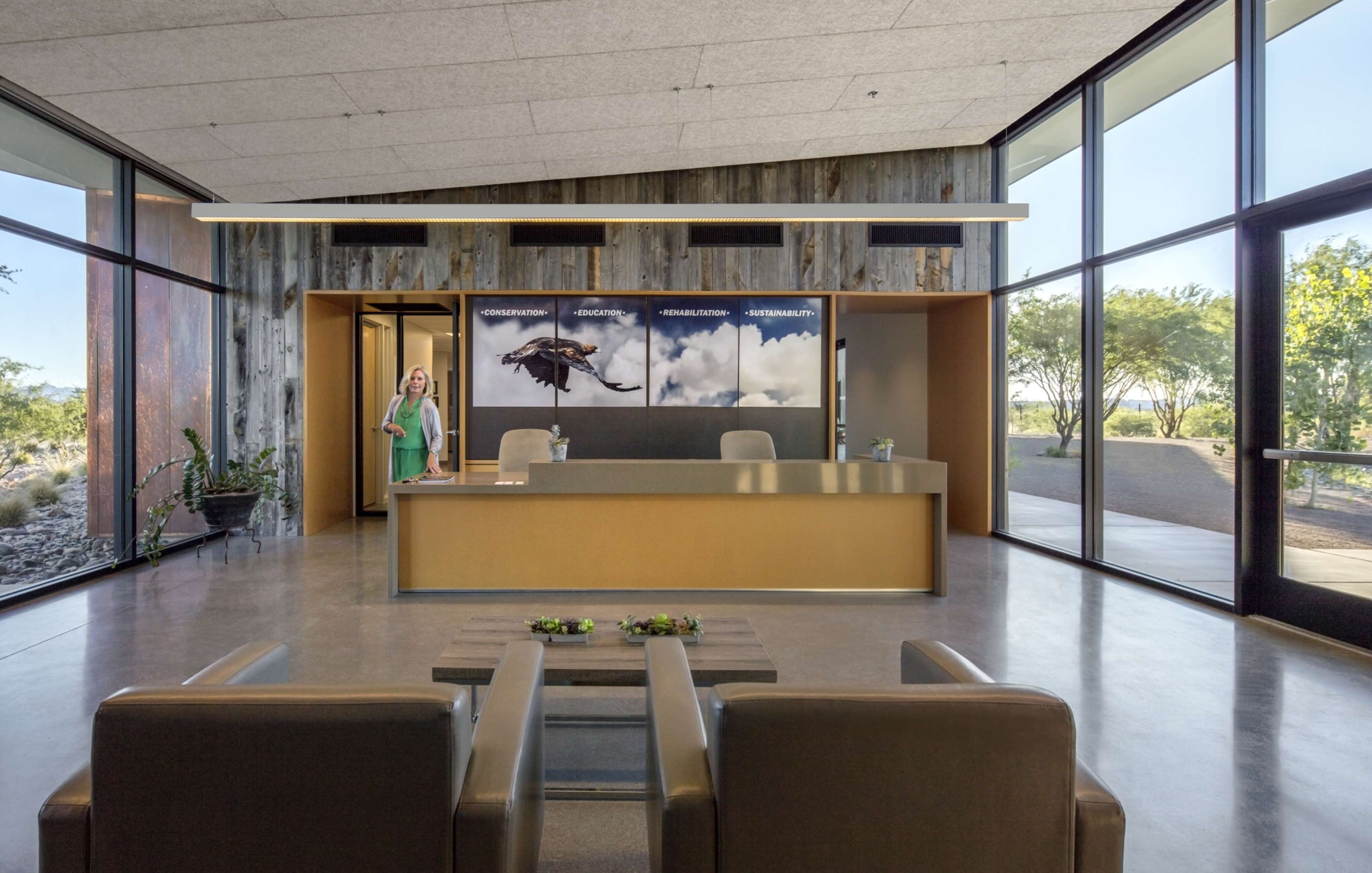 Modern office lobby with large windows, a central reception desk, and a person standing near the desk. The back wall displays three framed photographs, and there are seating arrangements in the foreground.