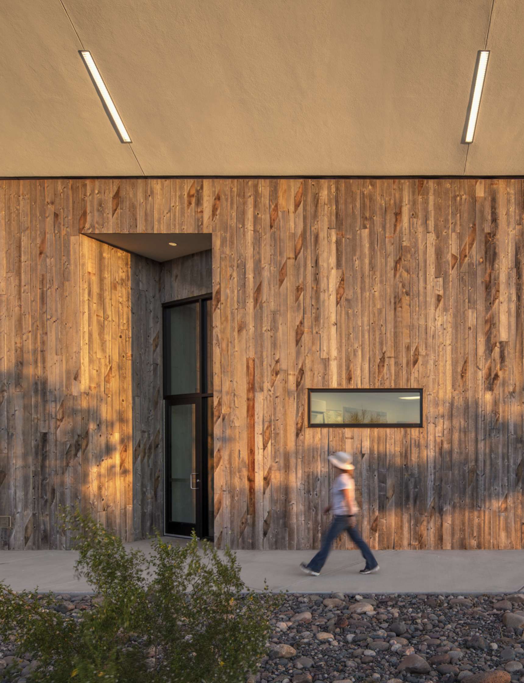 A person wearing a hat and jeans walks on a sidewalk in front of a wooden building with a large door and narrow window.