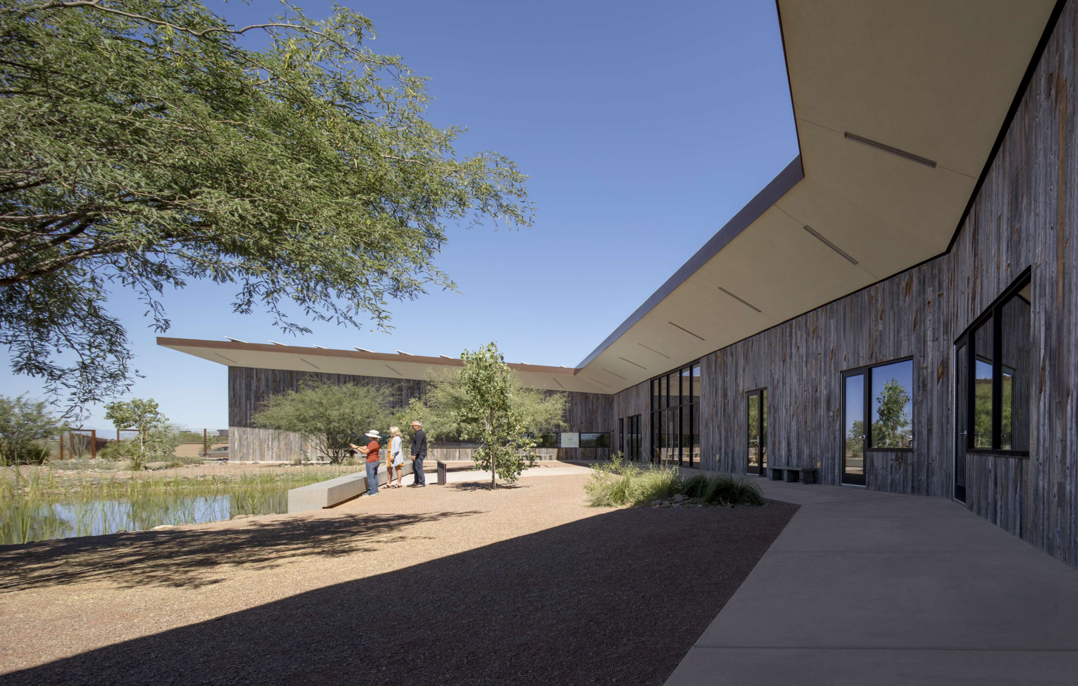 A modern building with a wooden exterior and angular roofline. A group of people is gathered outside near a tree and a body of water, surrounded by a dry, landscaped area under a clear sky.