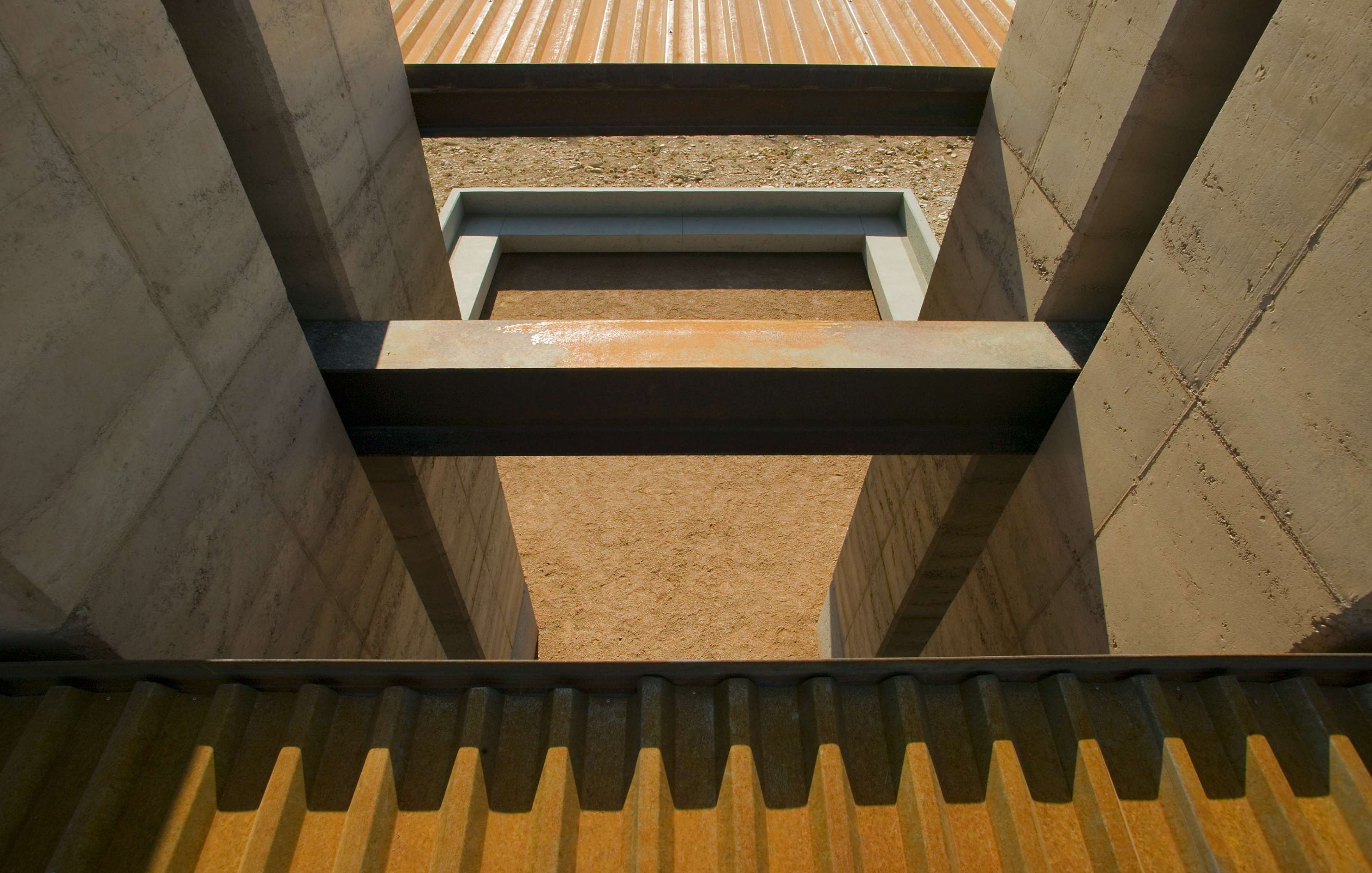 A view of a modern architectural structure with exposed concrete beams and corrugated metal surfaces arranged in linear patterns.