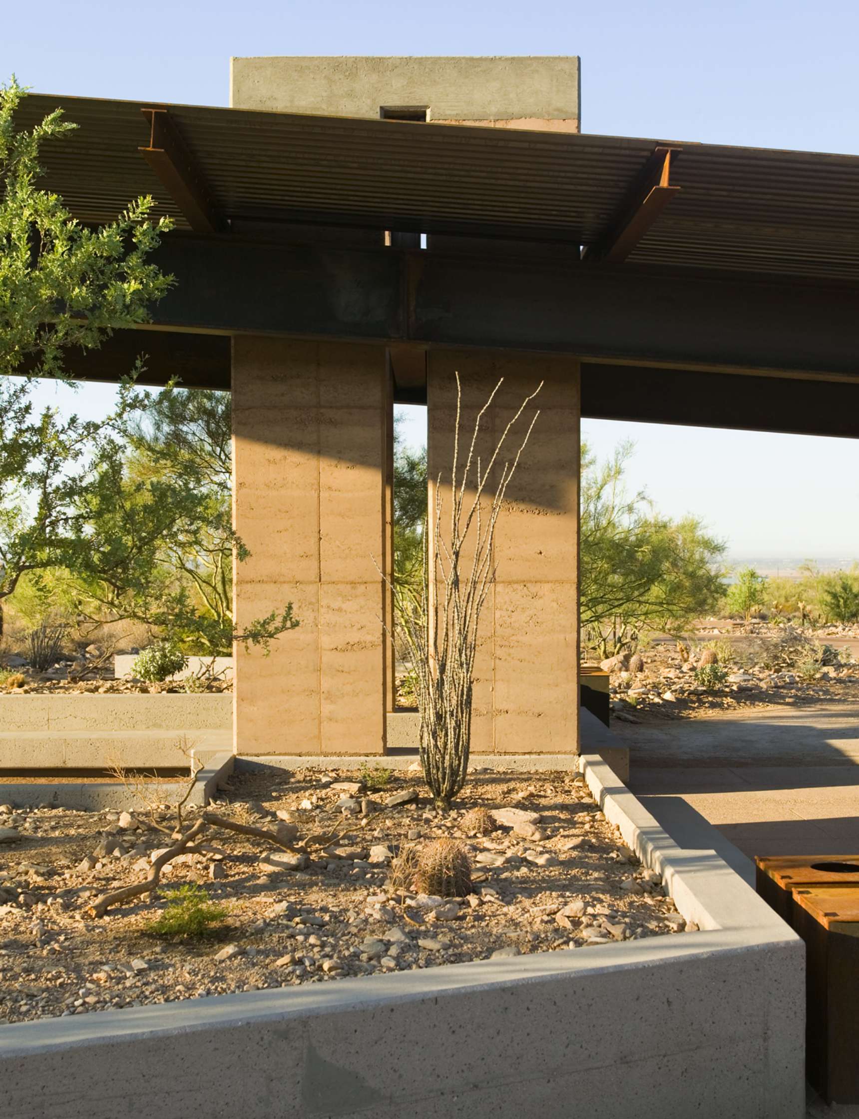 A modern building structure with concrete pillars and a desert landscape, featuring sparse vegetation including a tall, thin cactus.