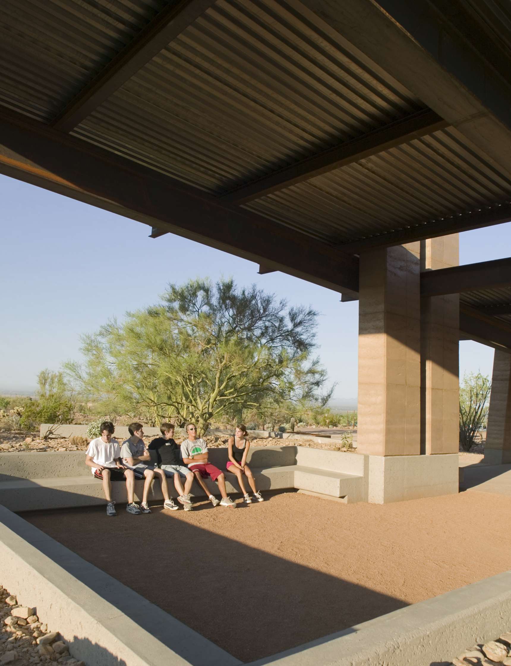 A group of five people sit on a shaded bench under a large wooden structure with desert scenery and a tree in the background.