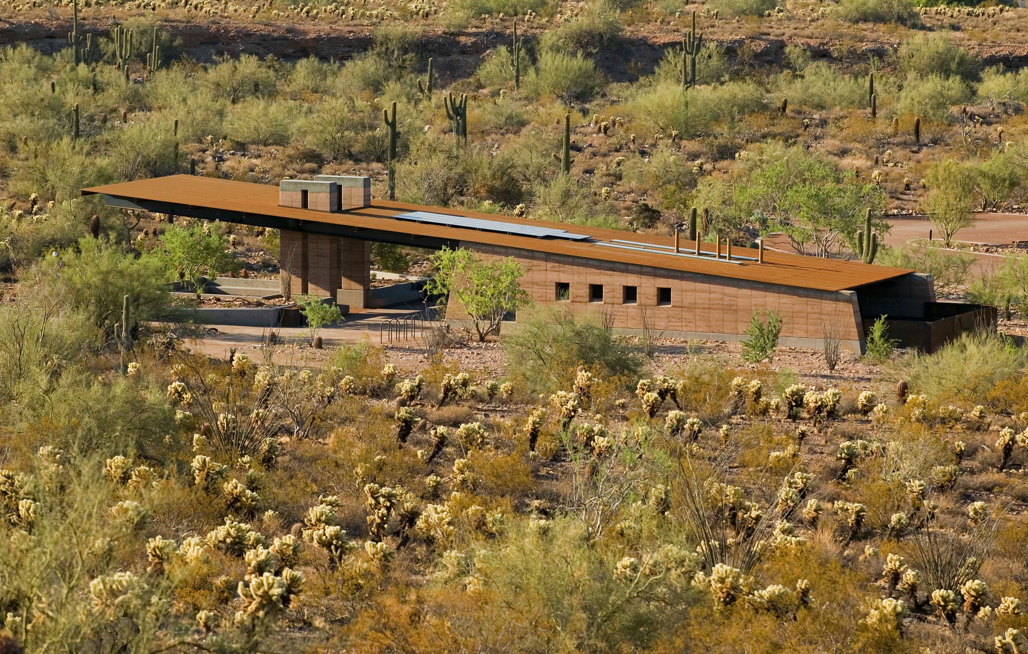 A modern, flat-roofed building situated in a desert landscape with sparse vegetation and cacti. The structure blends with the surroundings, featuring large, horizontal lines and earthy tones.