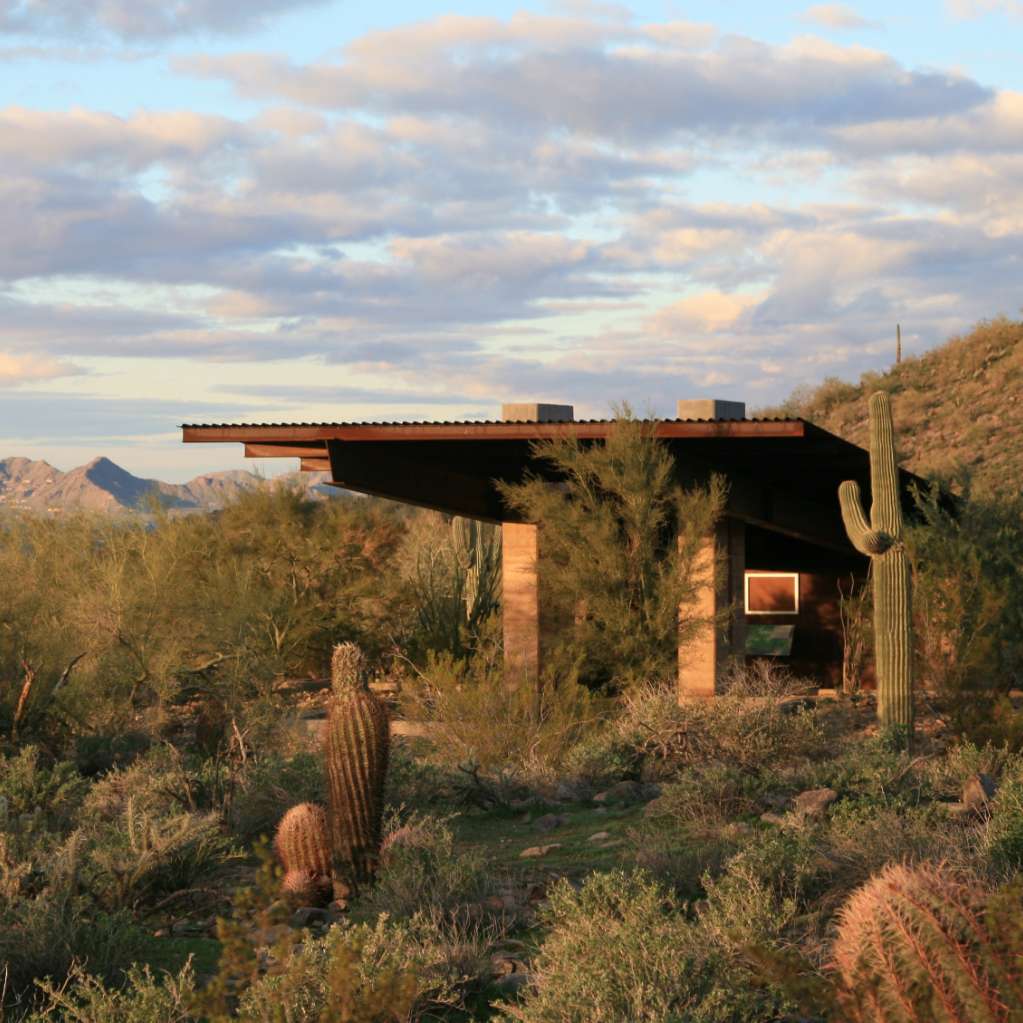 A small, modern house with a flat roof is nestled in a desert landscape with cacti and shrubs, set against a backdrop of mountains and a partly cloudy sky.