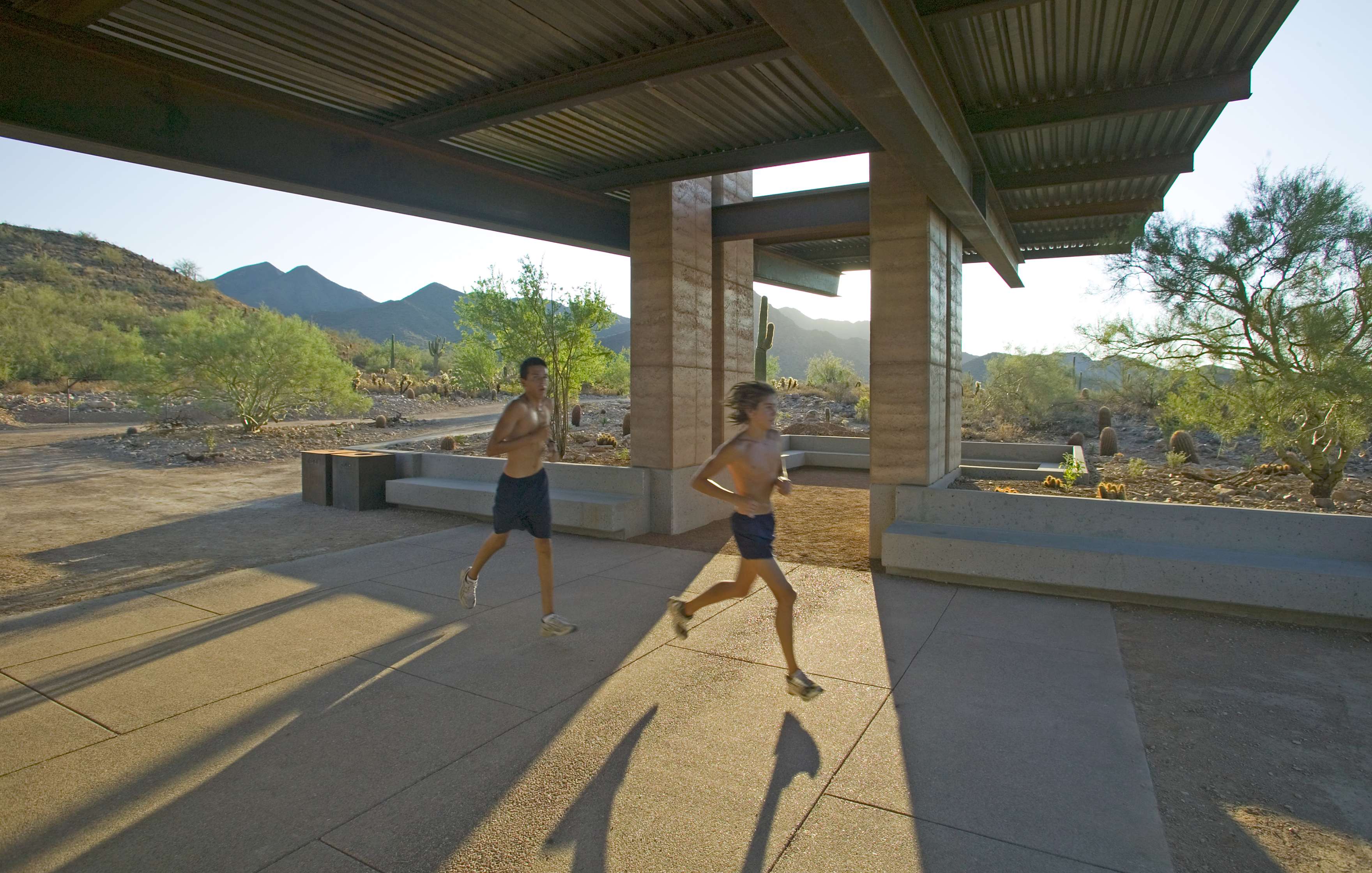 Two people running under a shaded structure in a desert landscape with mountainous terrain in the background.