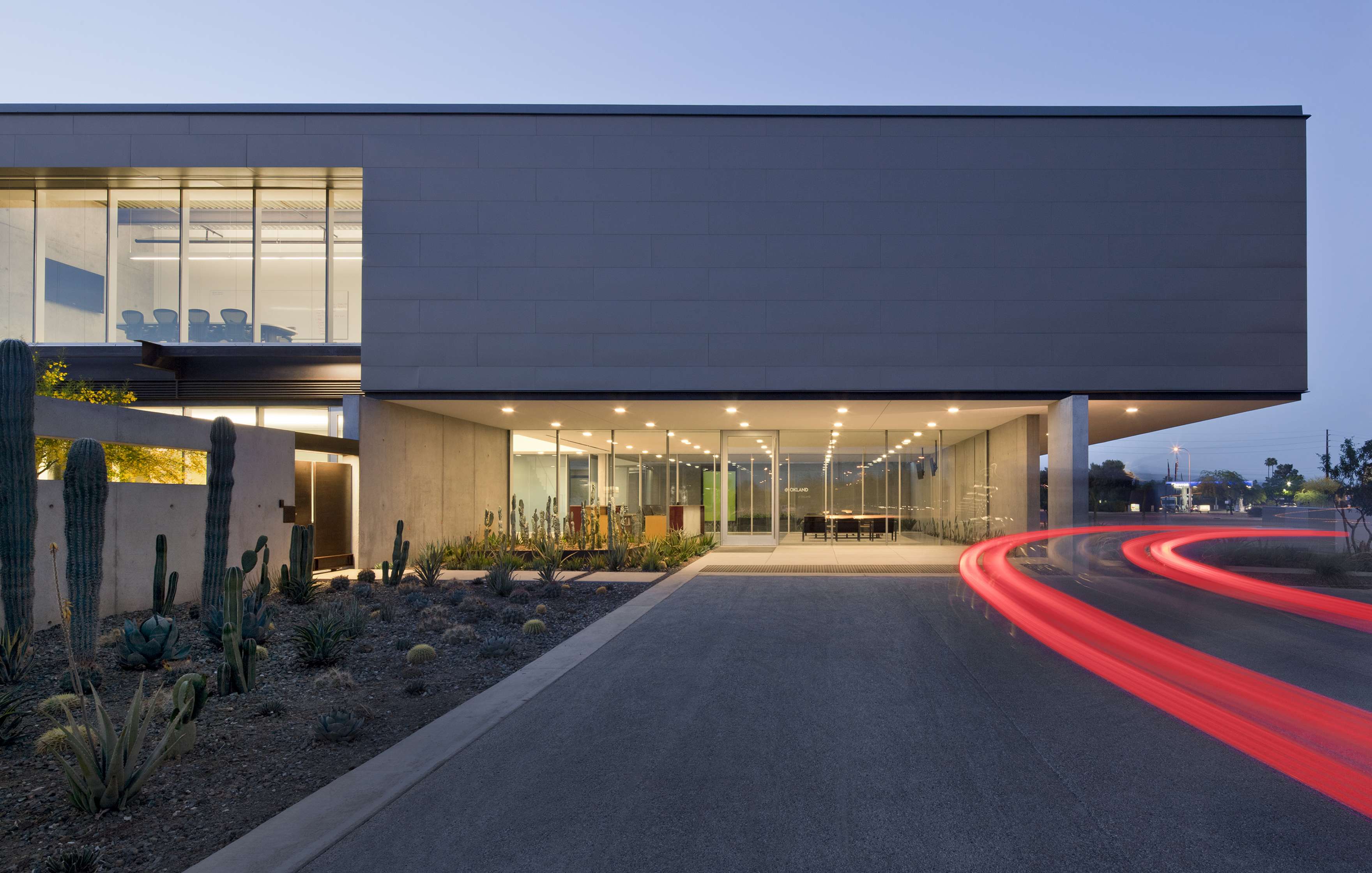 Modern building with large glass windows, illuminated at dusk. A streak of red light from a passing vehicle curves in front. Desert landscaping, including cacti, lines the sidewalk.