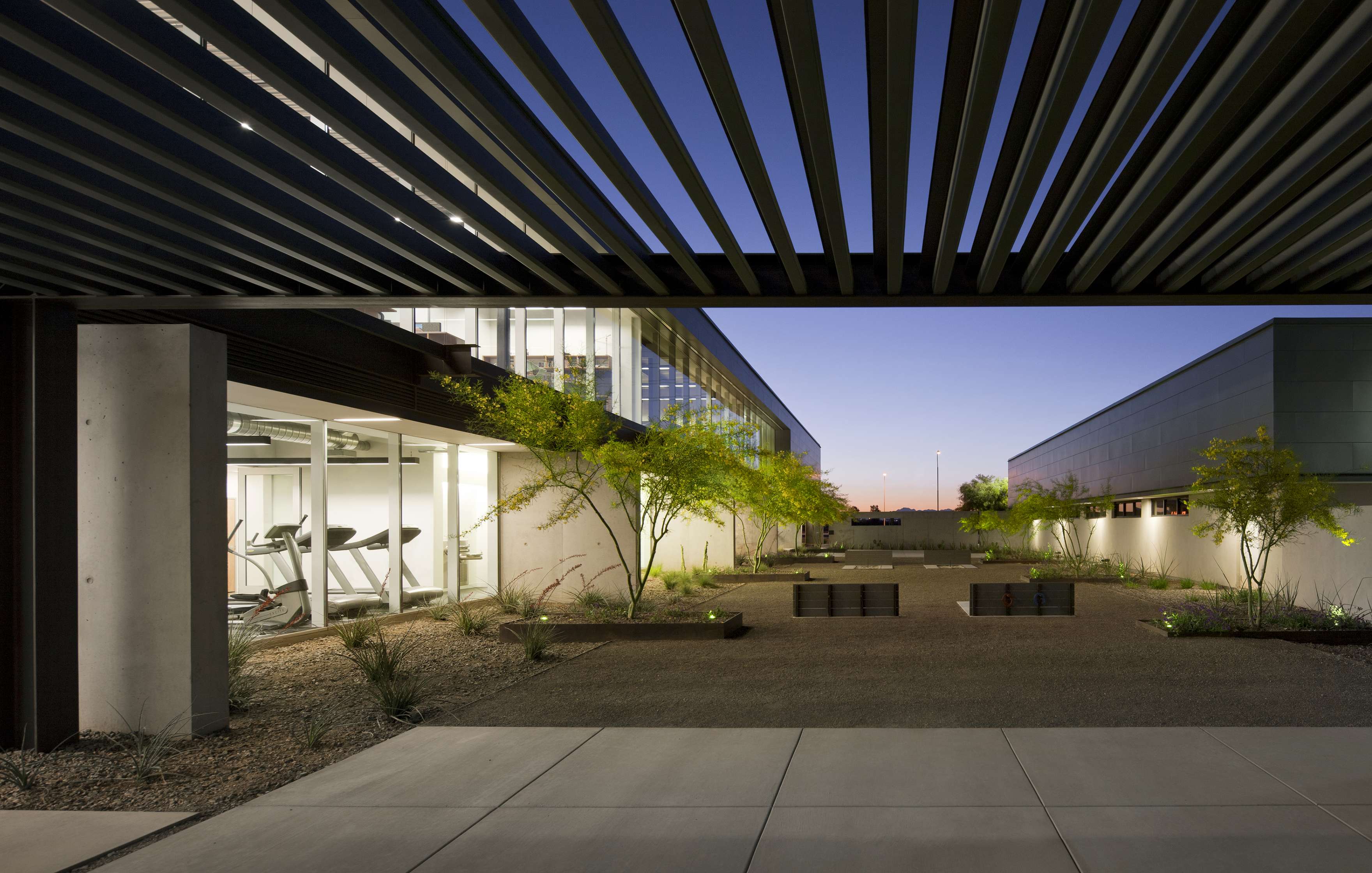 Modern building exterior with floor-to-ceiling windows revealing workout equipment inside. A spacious courtyard with minimalist landscaping is seen in the foreground under a louvered canopy.